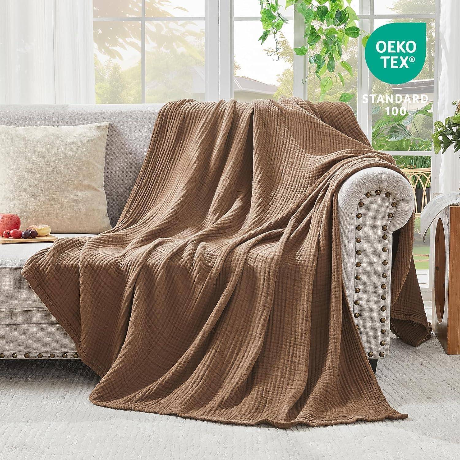 Eine gemütliche braune Decke, die über einem beigen Sofa in einem hellen Raum mit großen Fenstern und grünen Pflanzen liegt. Ein Etikett zeigt den Aufdruck 