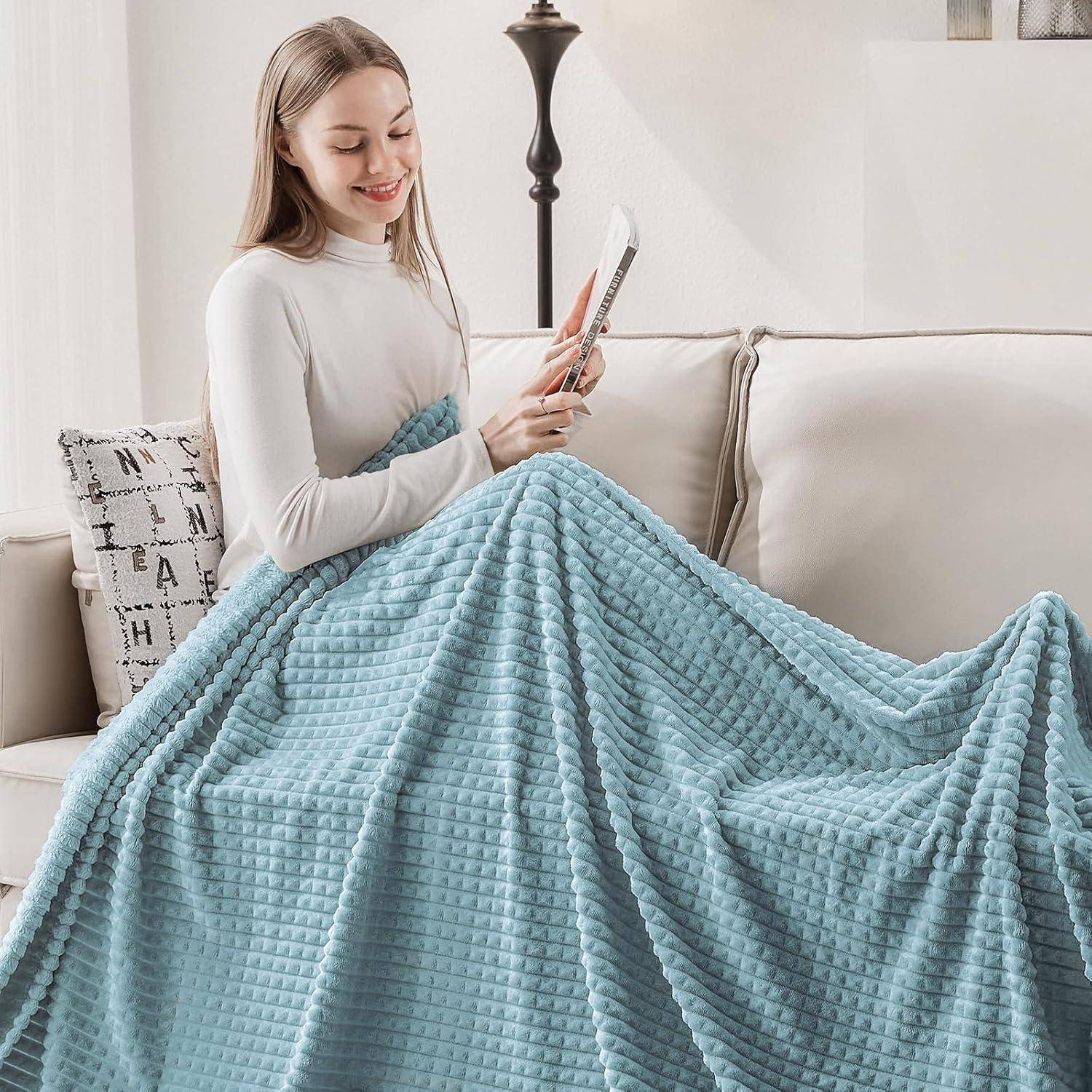 Eine lächelnde Person sitzt auf einem Sofa unter einer strukturierten blauen Decke und hält ein Mobiltelefon. Der Raum wirkt gemütlich und einladend.