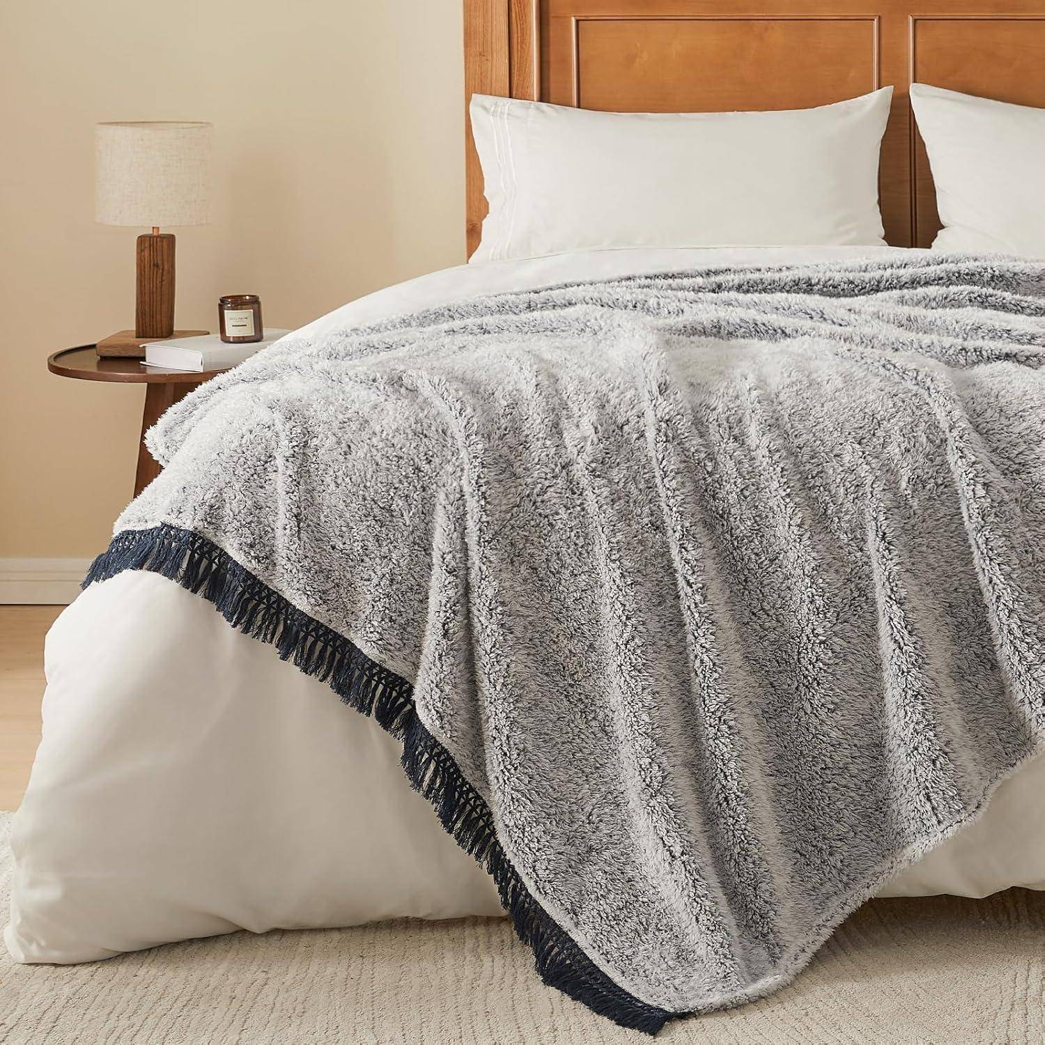 Ein ordentlich gemachtes Bett mit weißen Laken und Kissen, bedeckt mit einer strukturierten, grauen Decke mit einem dunkelblauen Fransenrand, aufgestellt in einem gemütlichen Schlafzimmer.