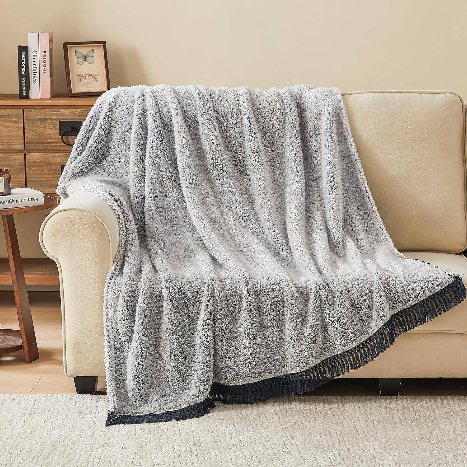 Eine flauschige graue Decke mit dunkler Franse, die über einem beigen Sofa drapiert liegt, neben einem Holzseitentisch mit Büchern und einer gerahmten Schmetterlingsillustration.