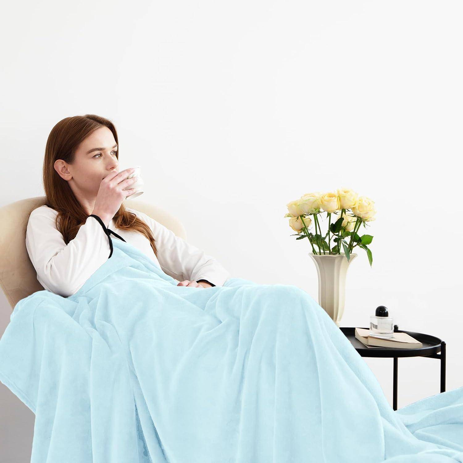 Eine Person, in eine hellblaue Decke gehüllt, sitzt auf einem Sofa und trinkt aus einer Tasse, während auf einem nahe gelegenen Tisch eine Vase mit gelben Blumen steht.