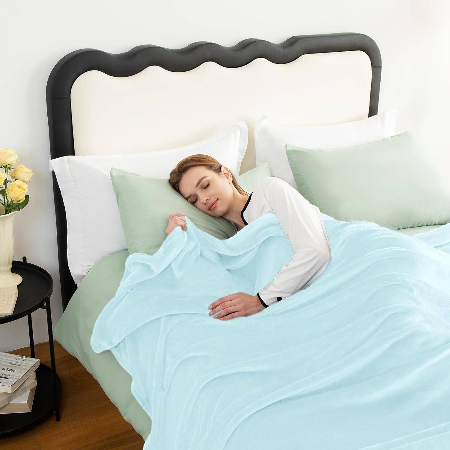Eine Frau schläft friedlich auf einem Bett mit hellblauer Bettwäsche und minzgrünen Kissen. Ein Nachttisch mit Blumen und Büchern steht in der Nähe.