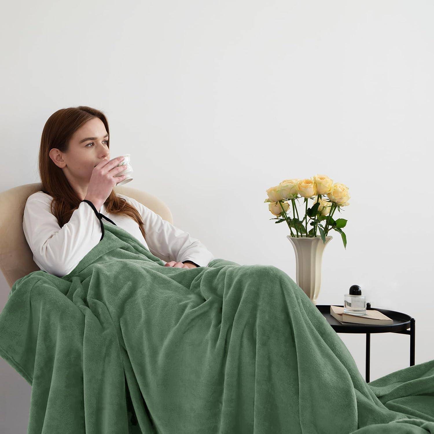 Eine Frau, in eine grüne Decke gehüllt, trinkt aus einer Tasse. Eine Vase mit gelben Rosen und ein Buch liegen auf dem Tisch neben ihr.