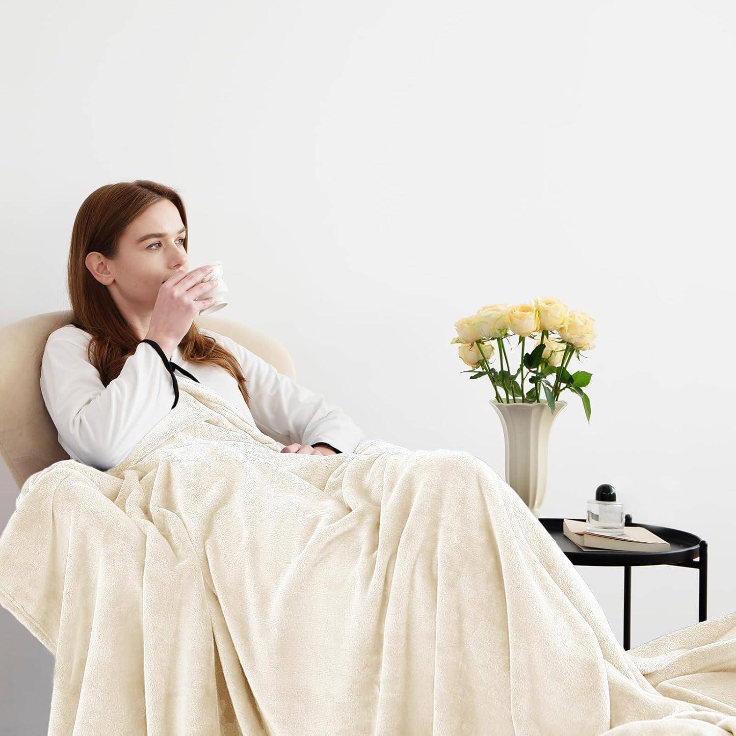 Eine Person sitzt auf einem Sofa, in eine cremefarbene Decke gehüllt, und nippt an einer Tasse. Eine Vase mit gelben Rosen und eine Kerze stehen auf einem Tisch in der Nähe.