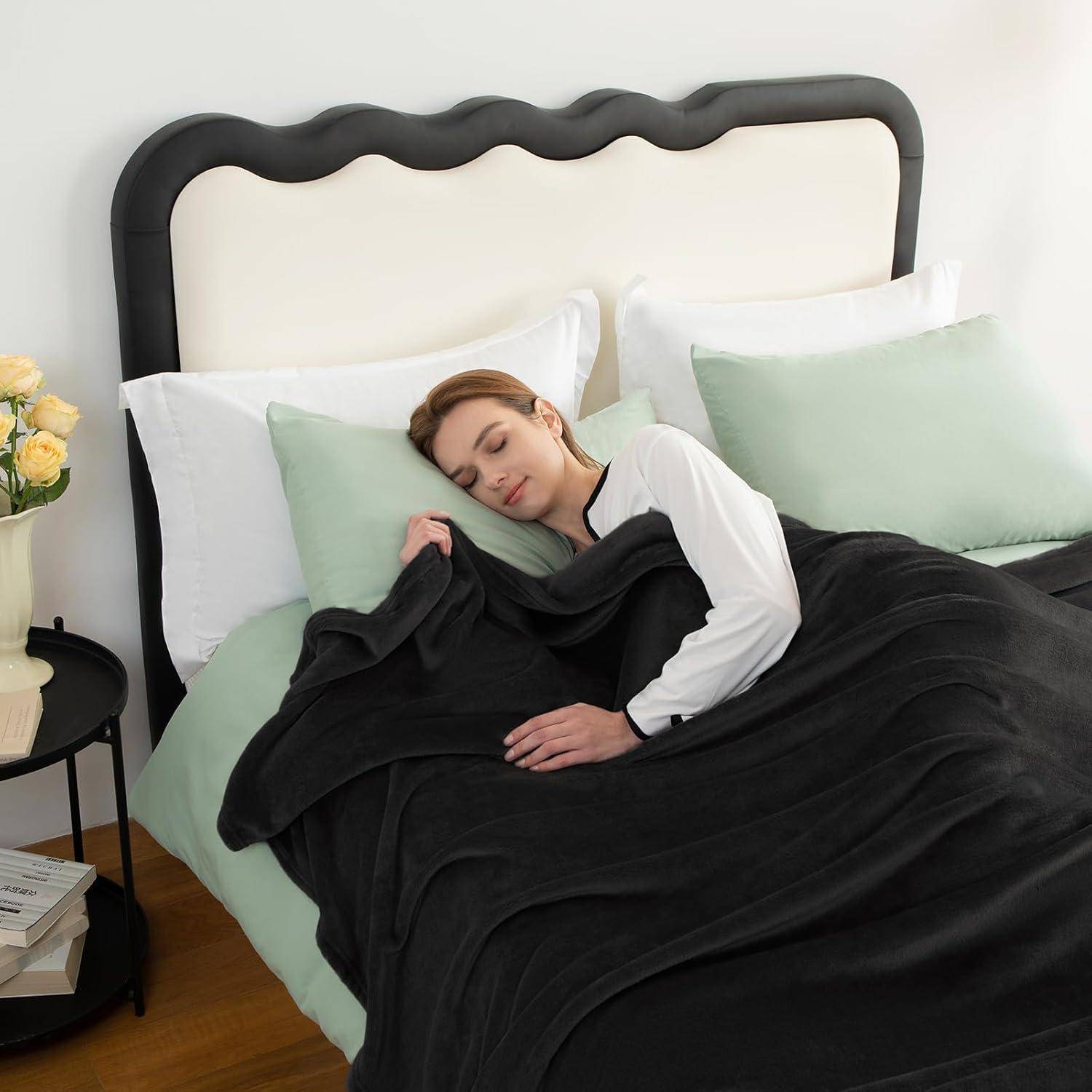 Eine Person schläft friedlich in einem sorgfältig gemachten Bett mit einer schwarzen Decke, weißen und grünen Kissen und einem schwarzen Kopfteil. Ein kleiner Tisch mit einem Buch steht in der Nähe.