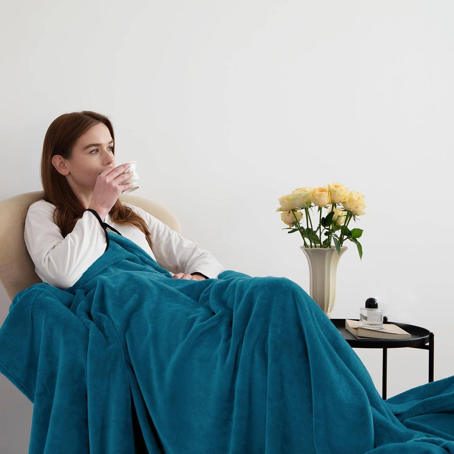 Eine Frau, in eine blaue Decke gehüllt, nippt an einem heißen Getränk. Eine Vase mit gelben Rosen und ein kleiner Tisch mit einem Telefon befinden sich neben ihr.