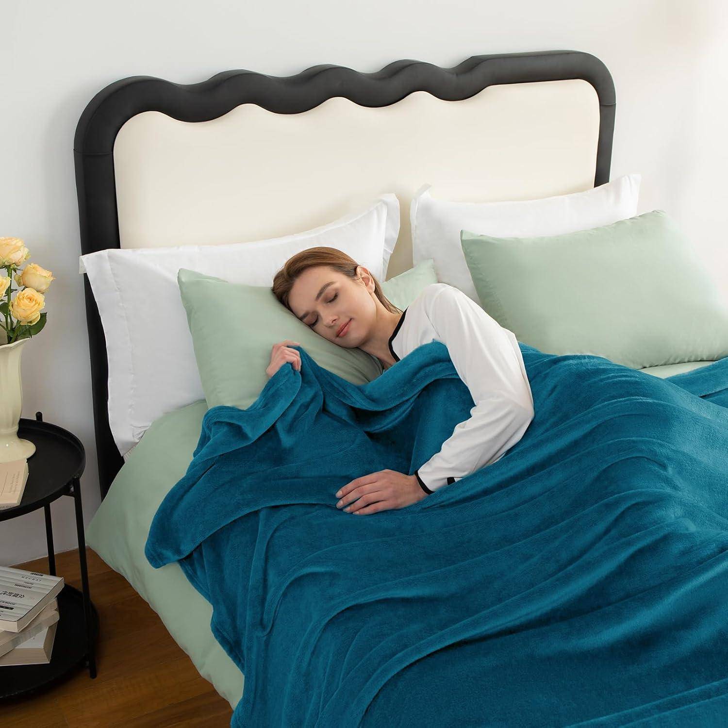 Eine Person schläft bequem auf einem Bett mit türkisfarbenen Decken, umgeben von weißen und blassgrünen Kissen. Ein kleiner Nachttisch trägt eine Lampe.
