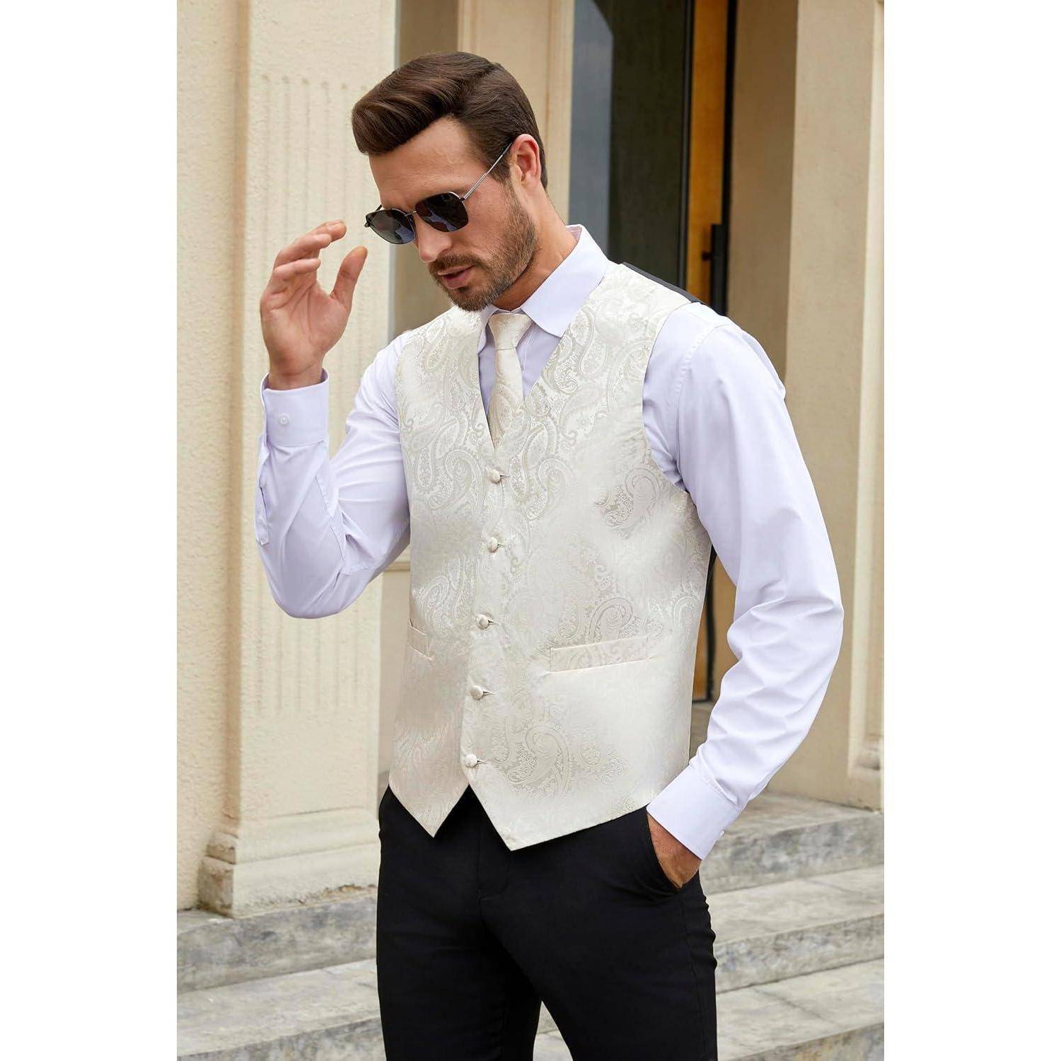 Ein Mann mit Sonnenbrille, der eine weiß gemusterte Weste über einem weißen Hemd trägt und lässig neben einem beigen Gebäude mit Stufen steht.