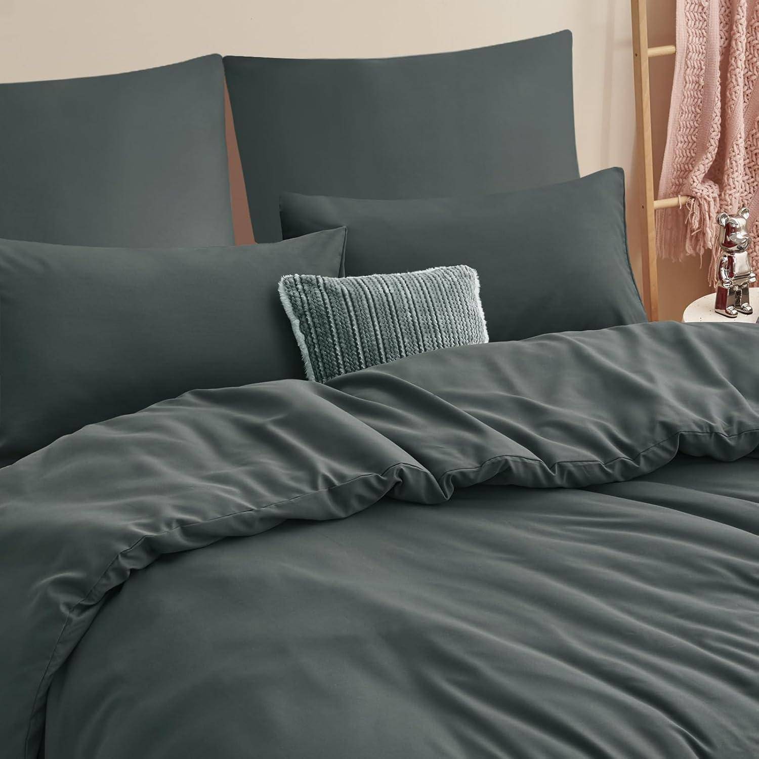 Ein ordentlich gemachtes Bett mit dunkelgrauer Bettwäsche und mehreren Kissen, darunter eines mit einer hellblauen gestrickten Kissenhülle, vor einem beigen Kopfteil.