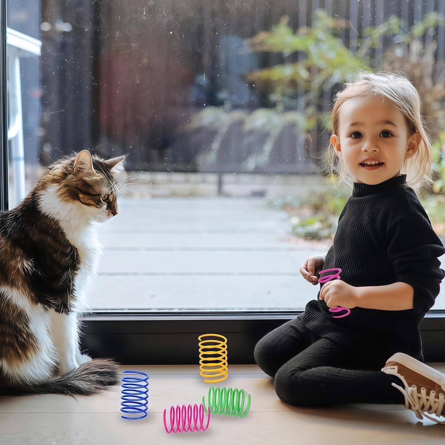Ein Kind sitzt am Fenster und lächelt, während es ein rosa Spielzeug hält. Eine Katze sitzt in der Nähe und blickt nach draußen. Bunte Spiralspielzeuge liegen auf dem Boden.