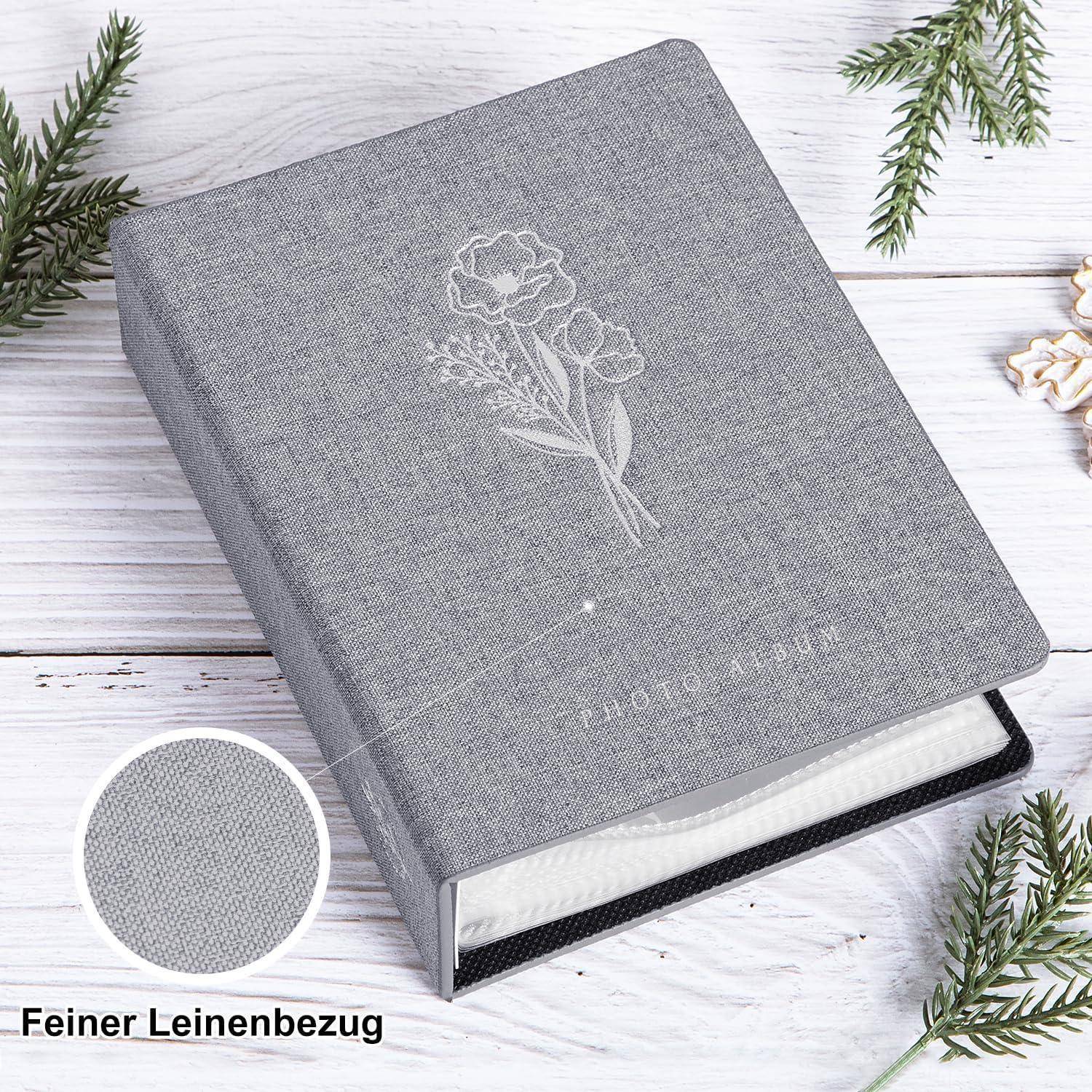 Ein graues Fotoalbum mit Blumenmuster und der Aufschrift 'Fotoalbum' auf dem Einband, umgeben von Tannenzweigen, das Details einer Stoffstruktur zeigt.
