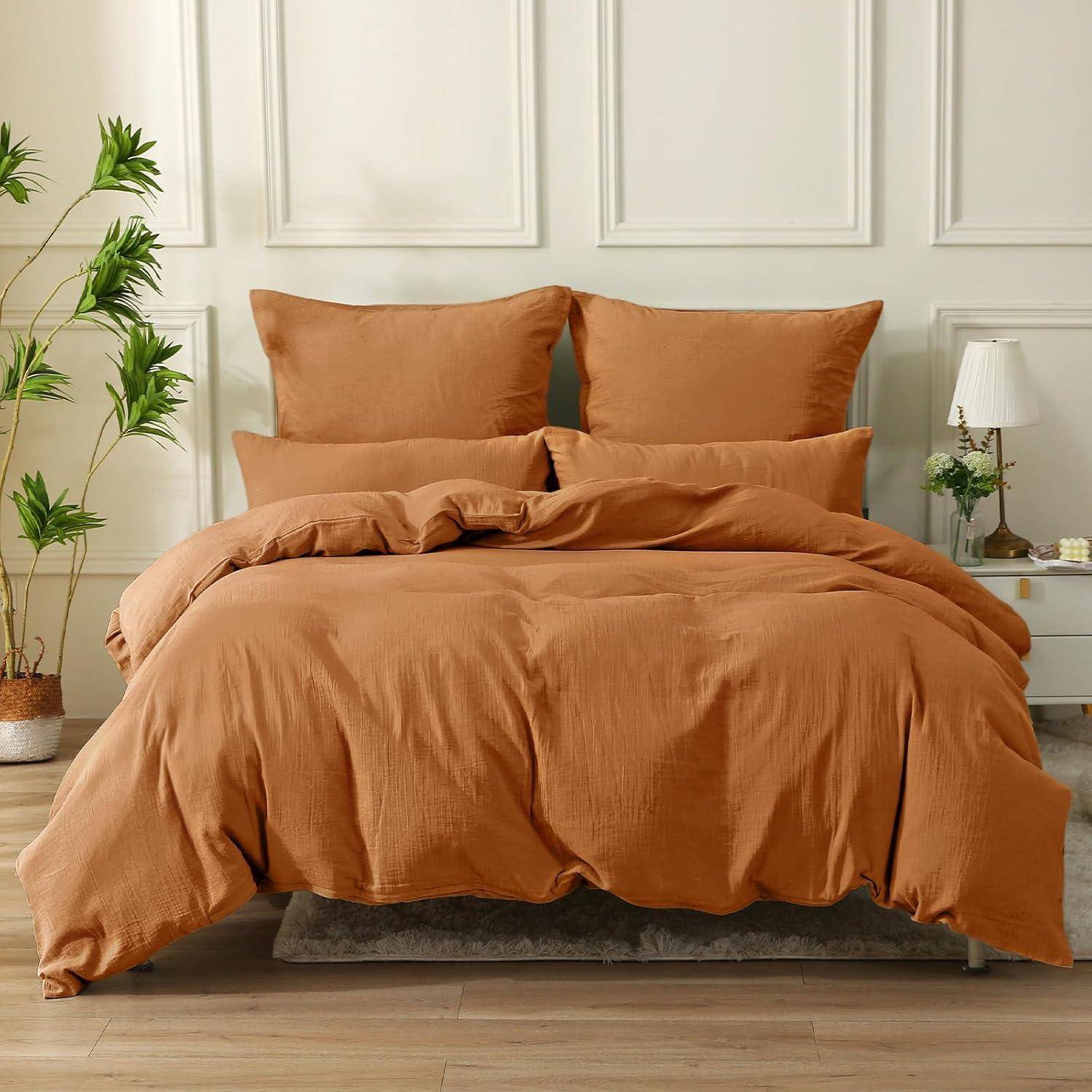 Ein ordentlich gemachtes Bett mit orangefarbenen Leintüchern und zwei Kissen. Ein kleiner weißer Nachttisch mit einer Lampe und Dekoration befindet sich rechts. Eine Pflanze steht links.