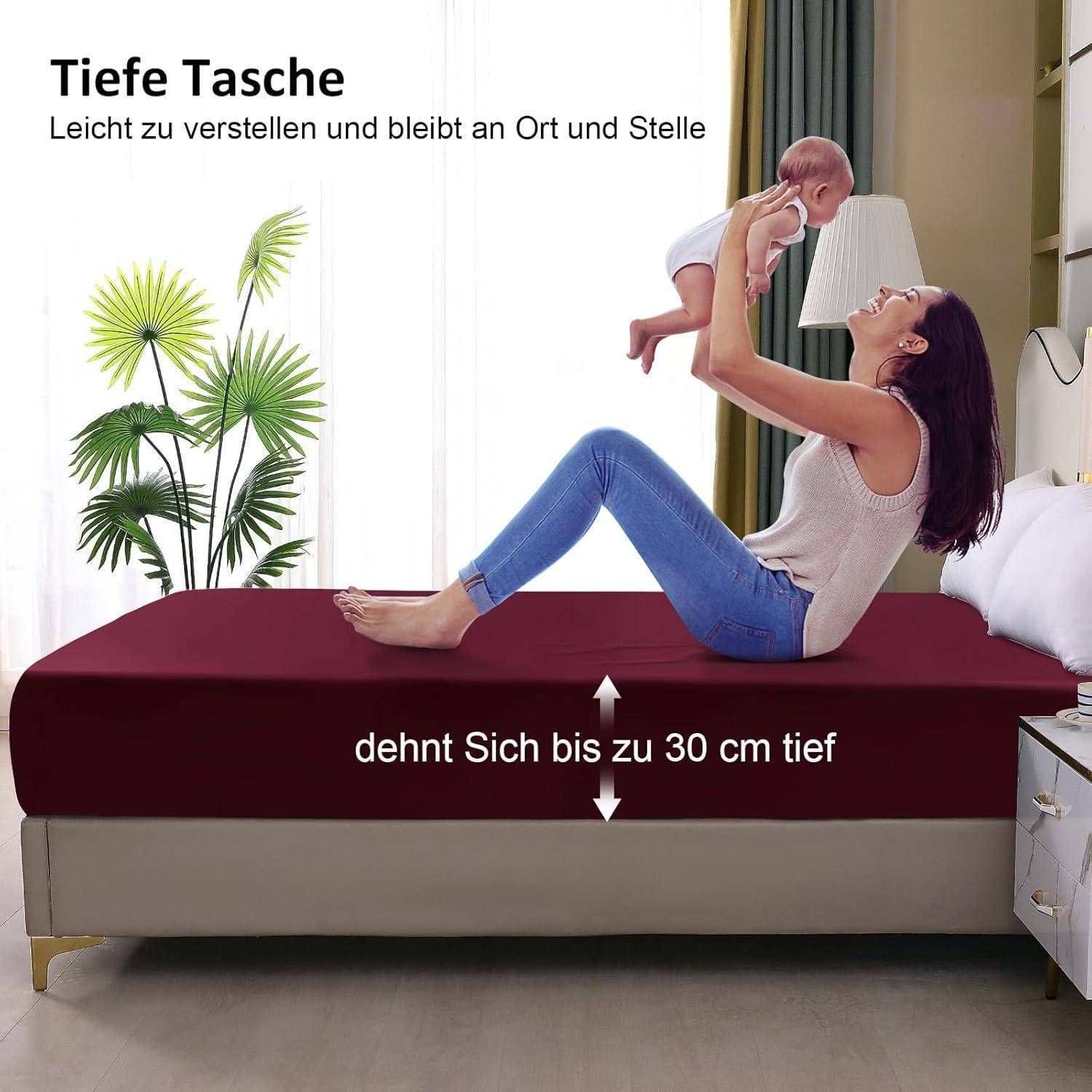 Eine Frau auf einem Bett hebt ein Baby in die Luft mit einem freudigen Ausdruck. Der Text beschreibt ein Spannbetttuch mit tiefen Taschen, das sich bis zu 30 cm anpassen lässt.