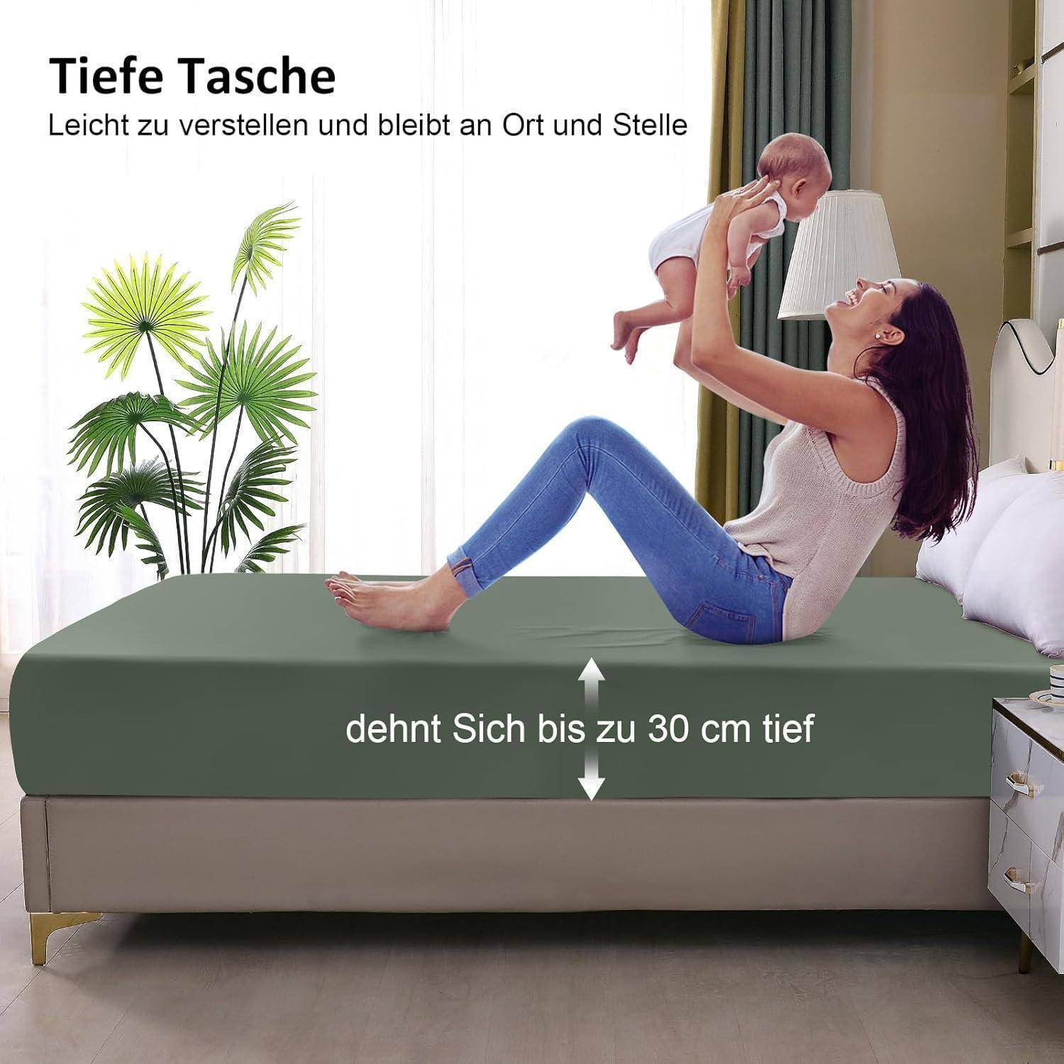 Eine Frau sitzt auf einem Bett und hebt ein Baby auf ihre Arme, interagiert fröhlich miteinander. Der Text auf Deutsch hebt den tiefen Dehnungsbereich des Bettlakens von bis zu 30 cm hervor.