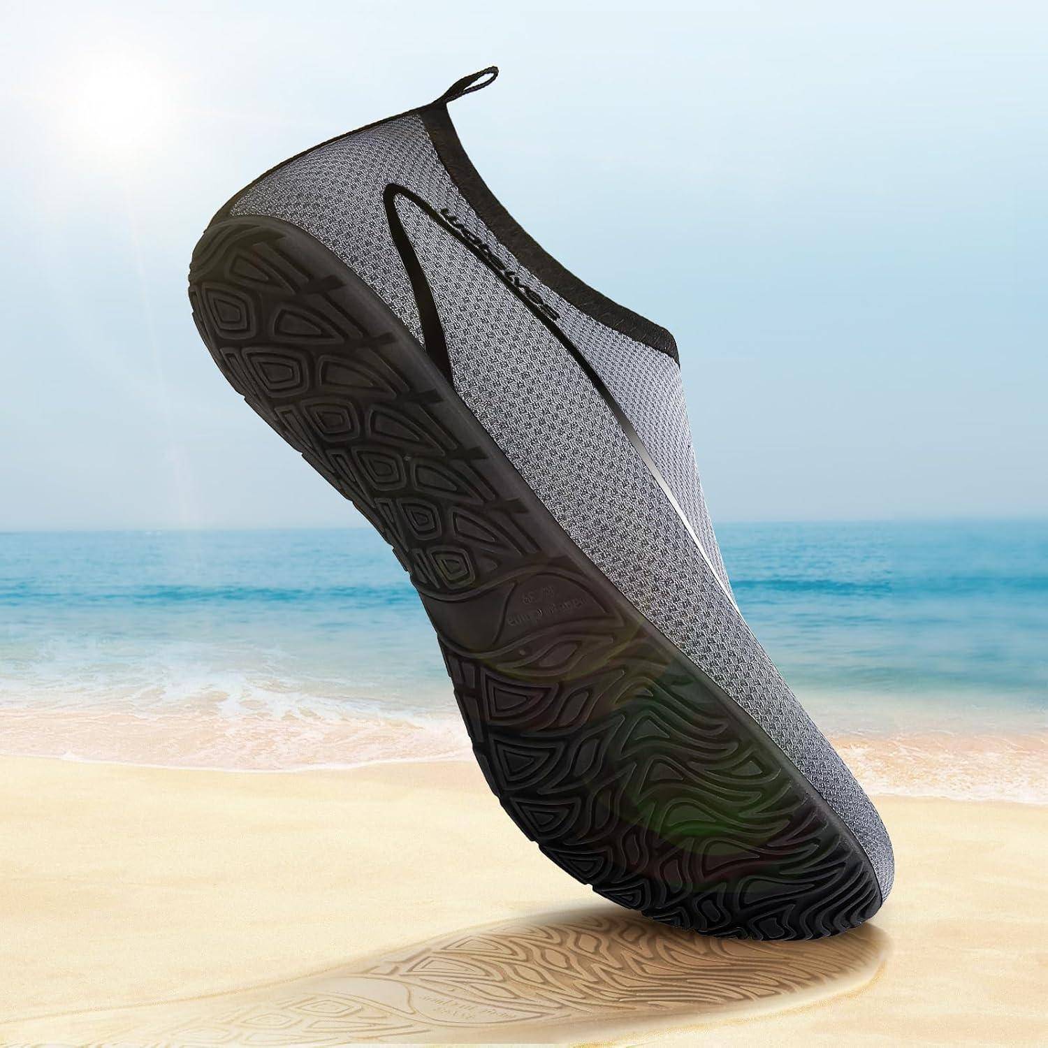Ein grauer Wasserschuh mit schwarzer Sohle befindet sich in der Sprungphase vor einem Strand-Hintergrund und unterstreicht dabei seine Flexibilität und sein Design.