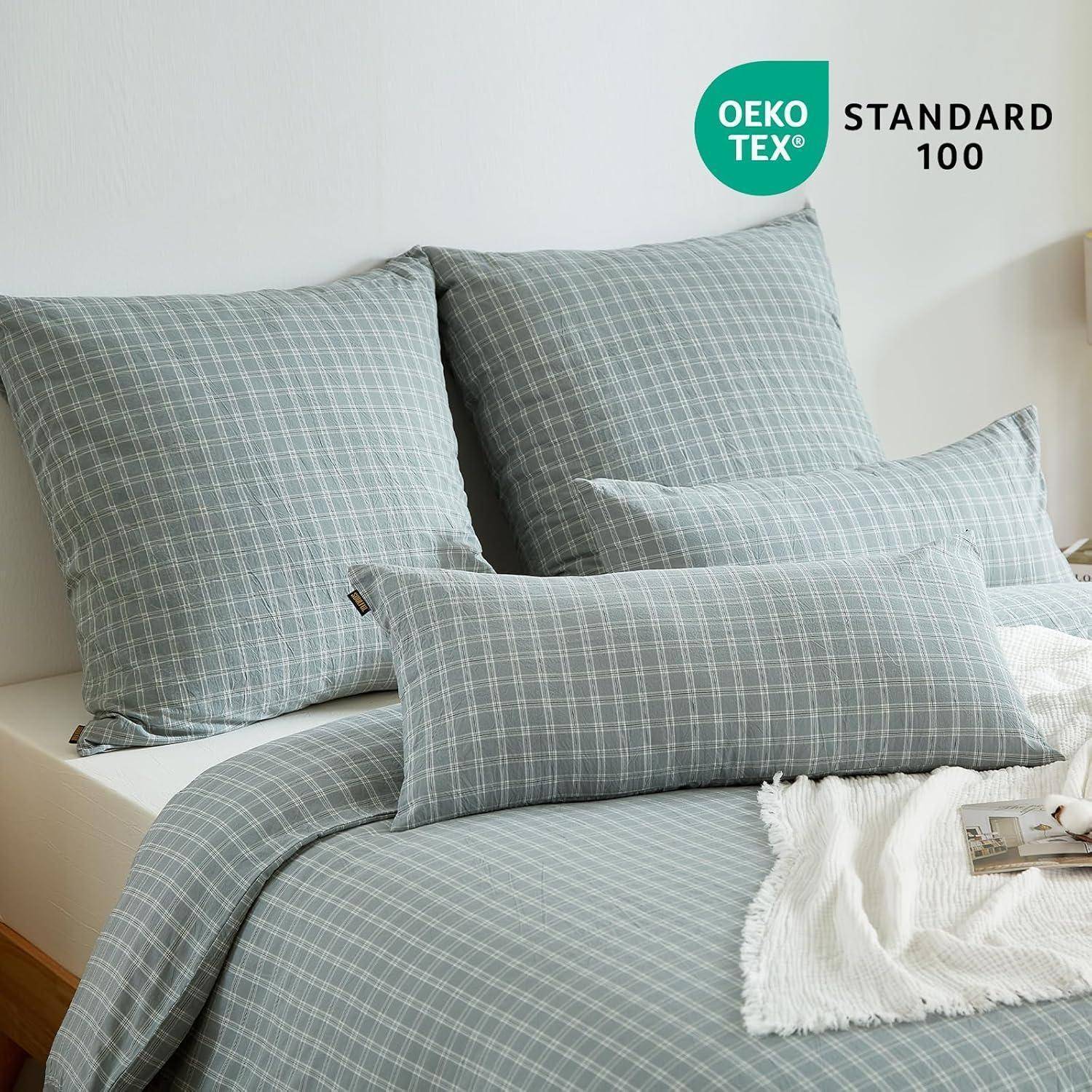 Ein Bett mit grau karierter Bettwäsche, einschließlich Kissen und Bettdecke, mit einer weißen, strukturierten Decke obenauf. 'OEKO-TEX STANDARD 100' Etikett.