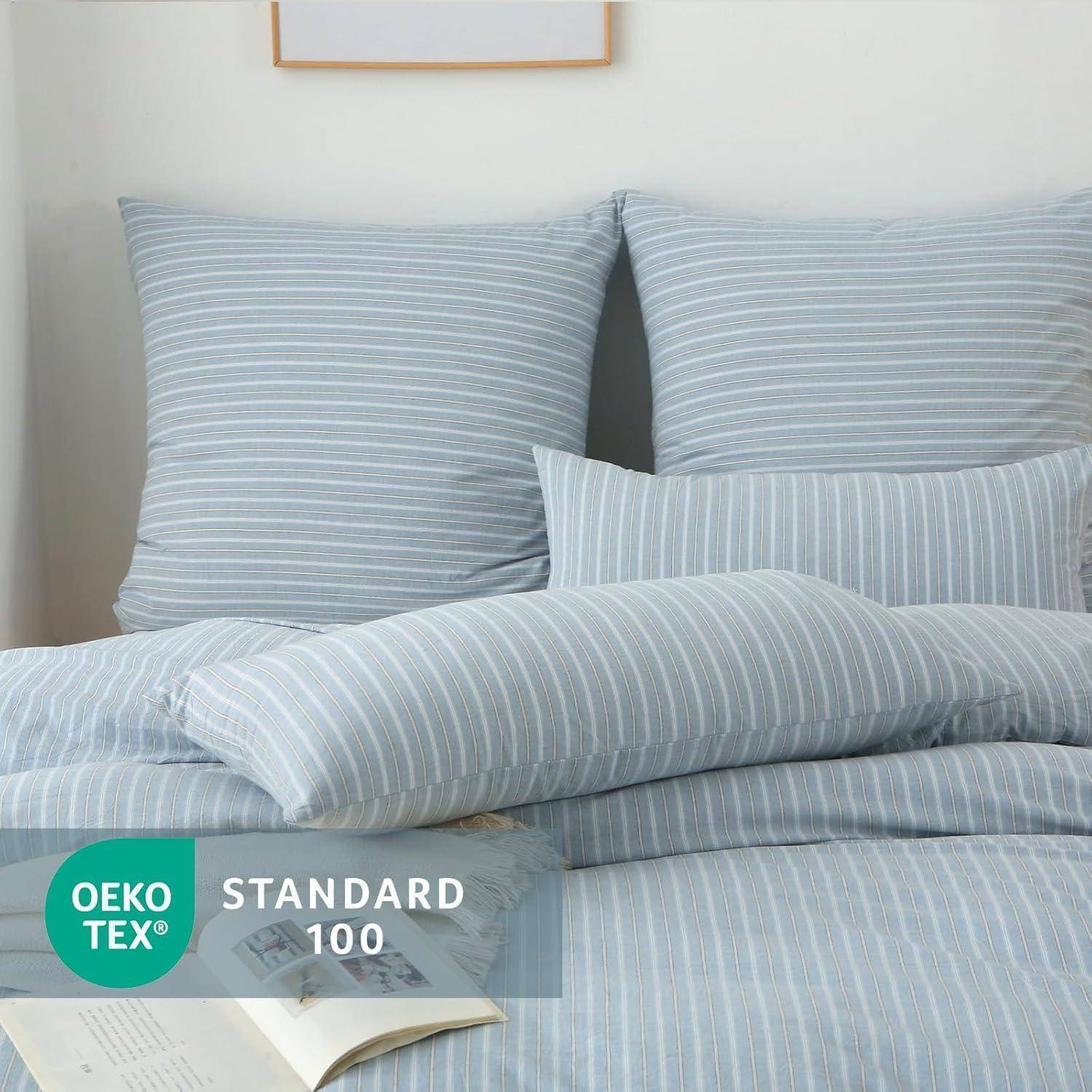 Bett mit hellgrauen und weißen gestreiften Kissen und Bettwäsche. 'OEKO-TEX Standard 100' Etikett am unteren Rand, das die Sicherheitszertifizierung des Gewebes anzeigt.
