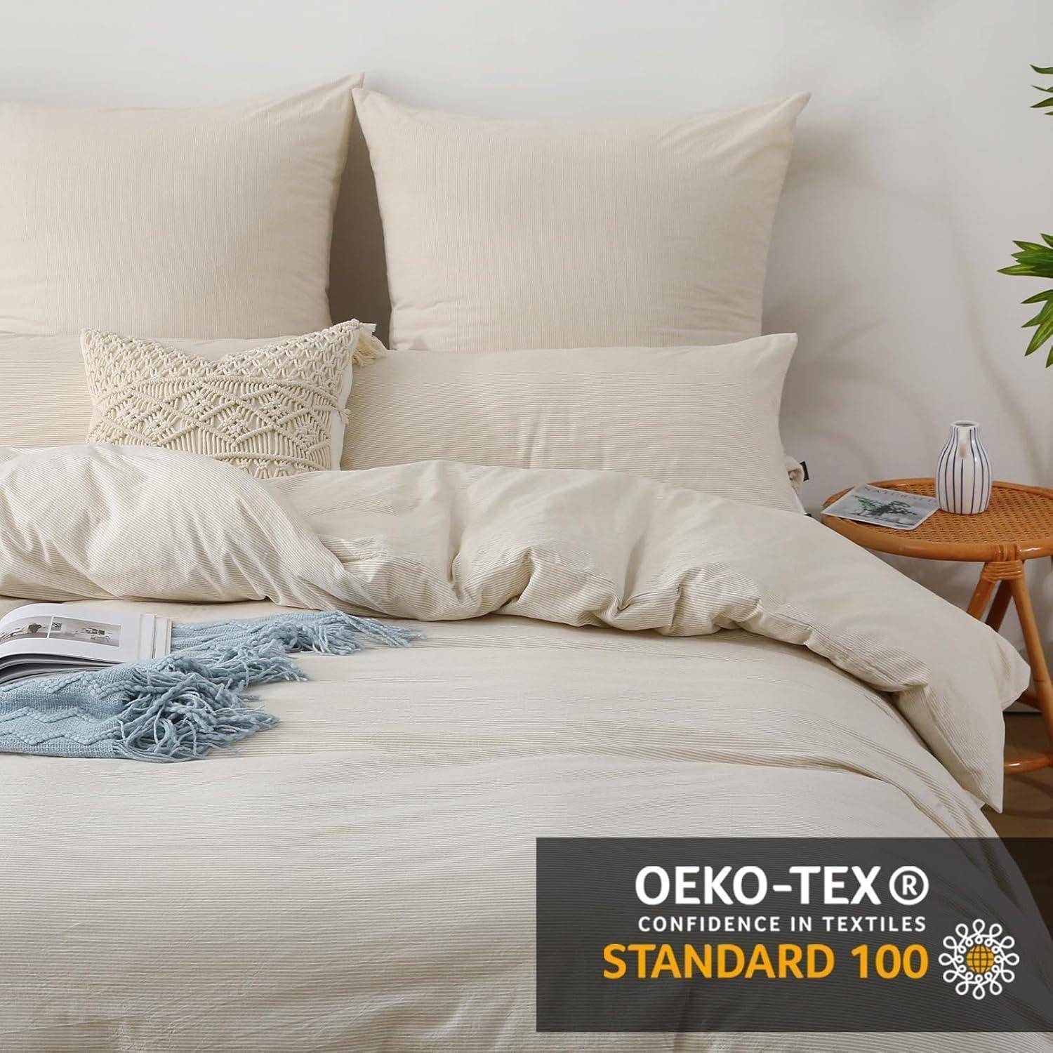 Ein sorgfältig gemachtes Bett mit cremefarbenen Bettwäsche und Kissen, einem geometrischen Dekokissen, einem Beistelltisch mit einer Pflanze und einem Buch, mit der Aufschrift 'OEKO-TEX Standard 100'.