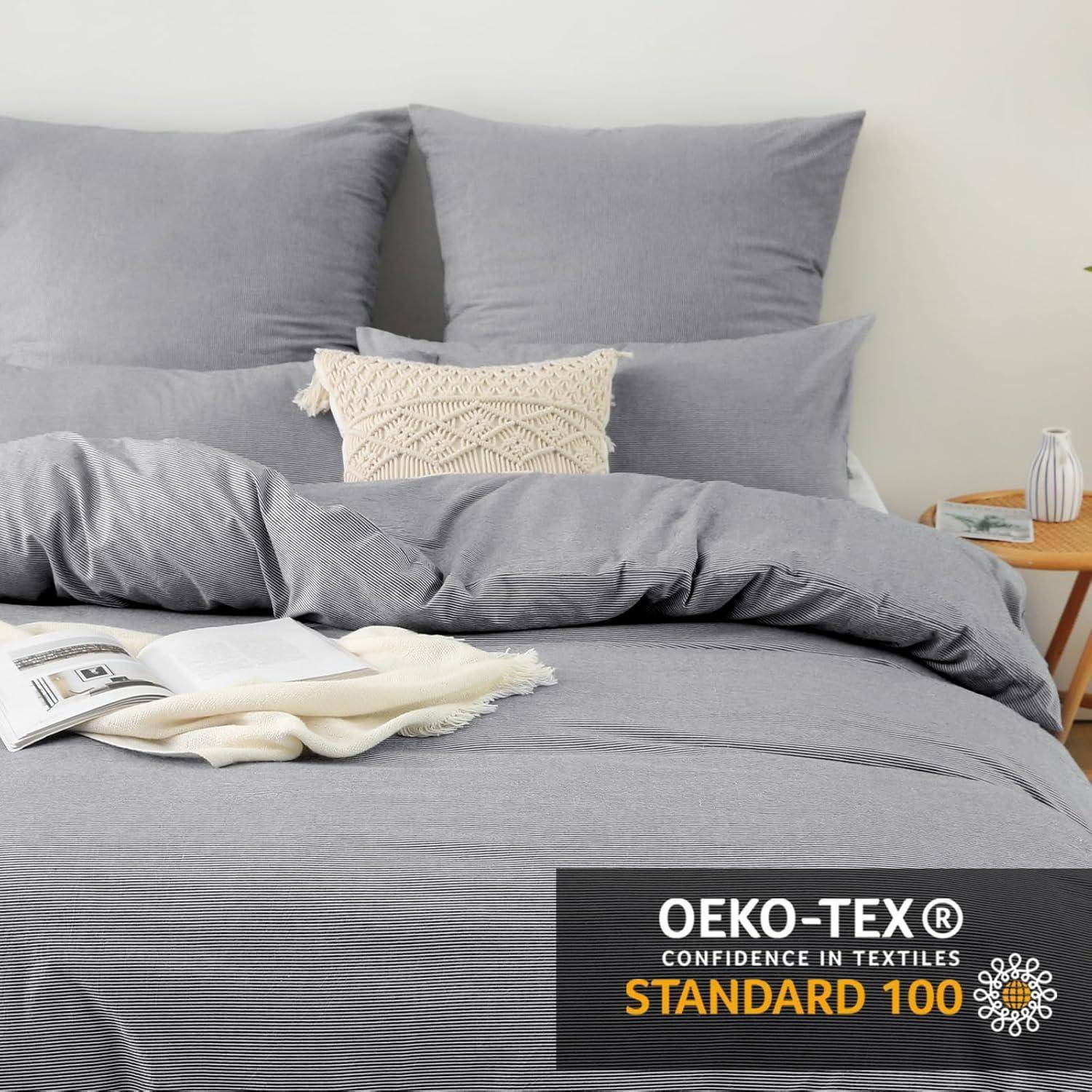 Ein sauber gemachtes Bett mit grauer Bettwäsche, dekorativen Kissen und einem Nachttisch mit einer Pflanze. Inklusive der 'OEKO-TEX STANDARD 100' Zertifizierung.