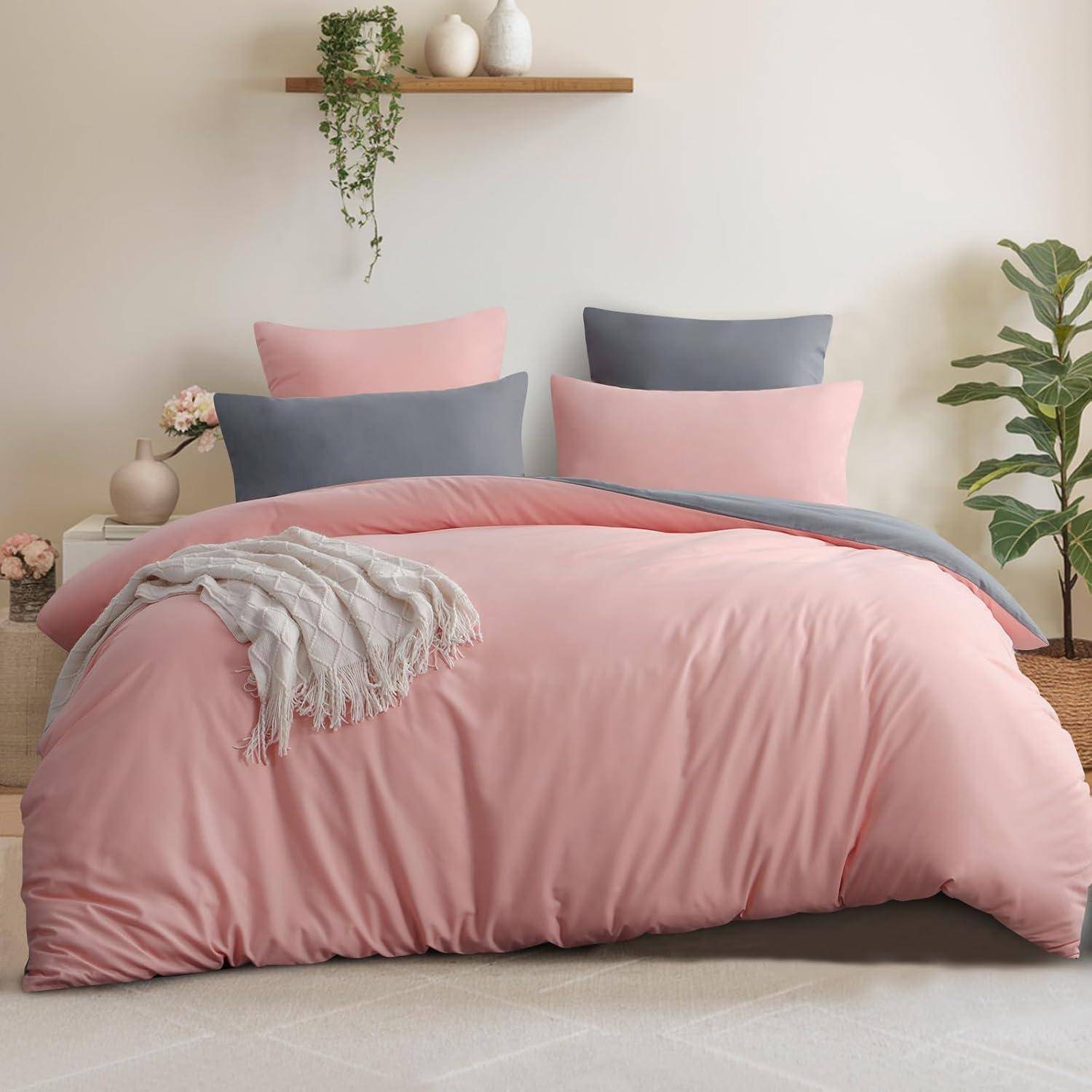 Ein ordentlich gemachtes Bett mit einer rosa Bettdecke, grauen und rosa Kissen und einem weißen Überwurf. Pflanzen und schlichte Dekorationen befinden sich in der Nähe.