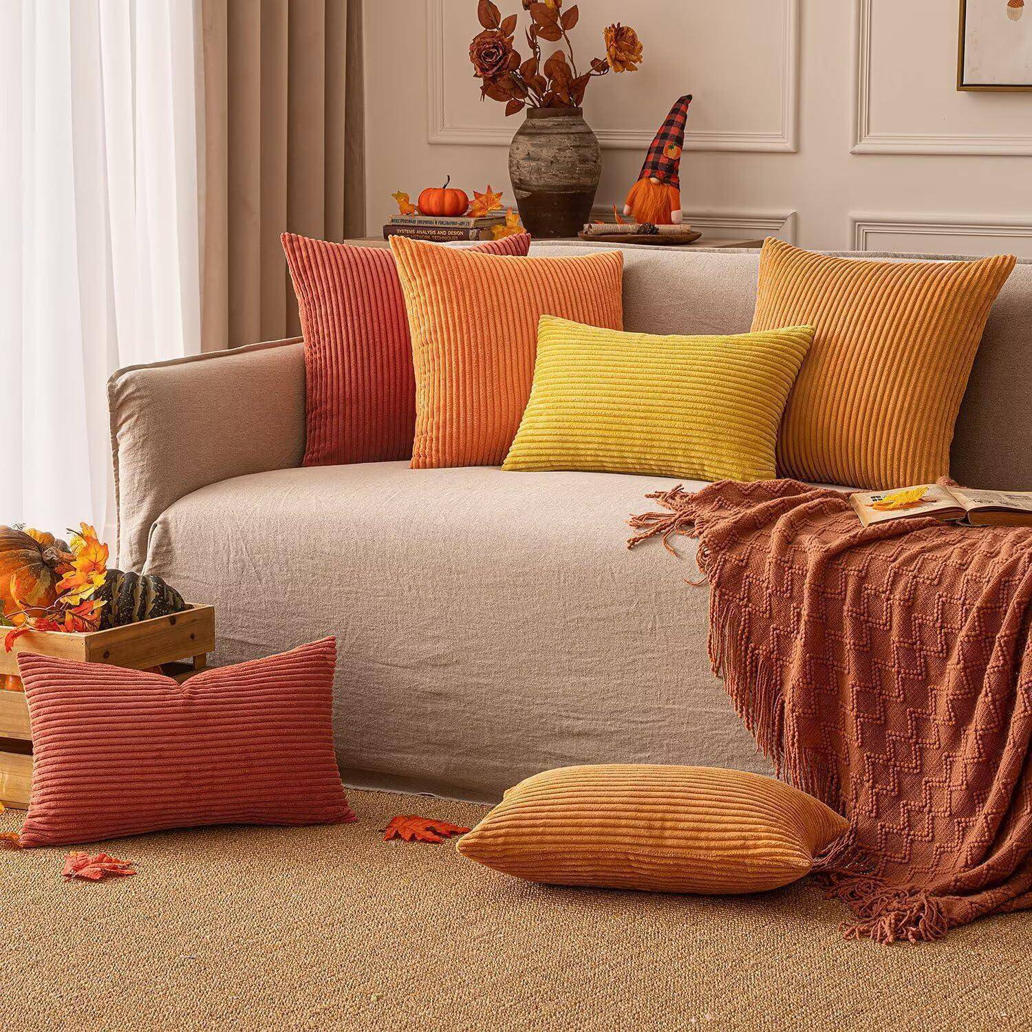 Ein gemütliches Wohnzimmer zeigt ein beiges Sofa mit orange-, gelben- und braunen Kissen, Herbst-inspirierter Dekoration und einer warmen Decke.