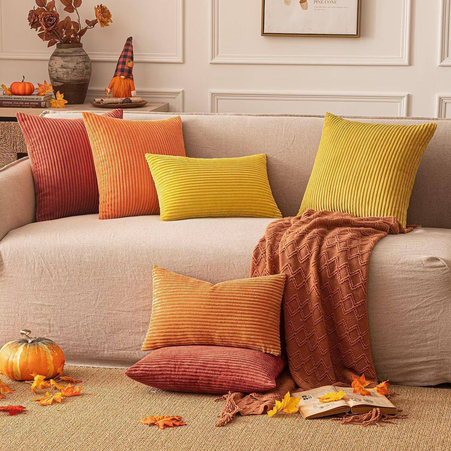 Eine gemütliche beige Couch mit bunten Zierkissen und einer gestrickten Decke in einem warmen, herbstlich gestalteten Wohnzimmer mit verstreuten Blättern.