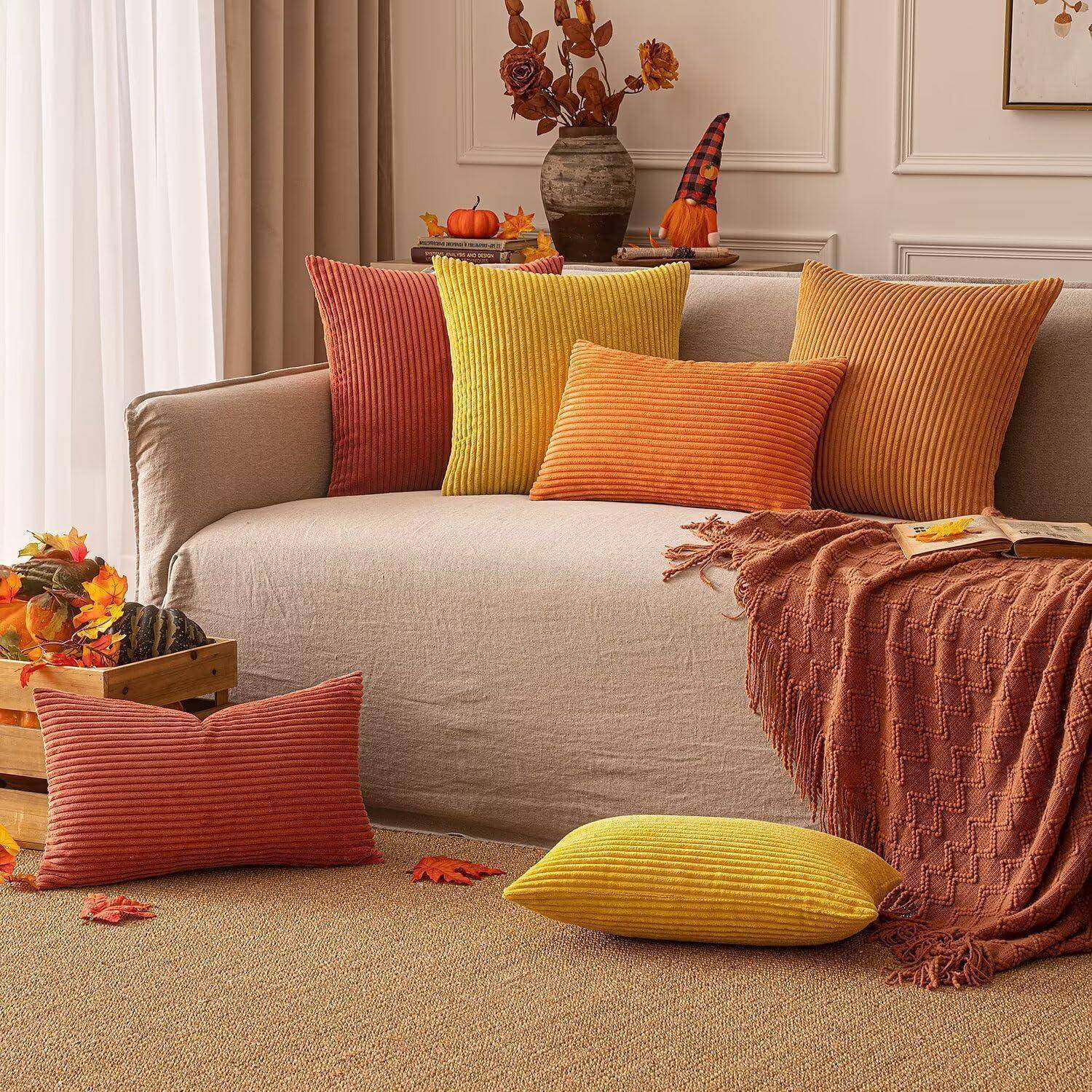Wohnzimmer mit einem beigen Sofa, akzentuiert durch rote, gelbe und orangefarbene Kissen und eine Decke. Herbstliche Dekoration umfasst Blätter und eine Vase.