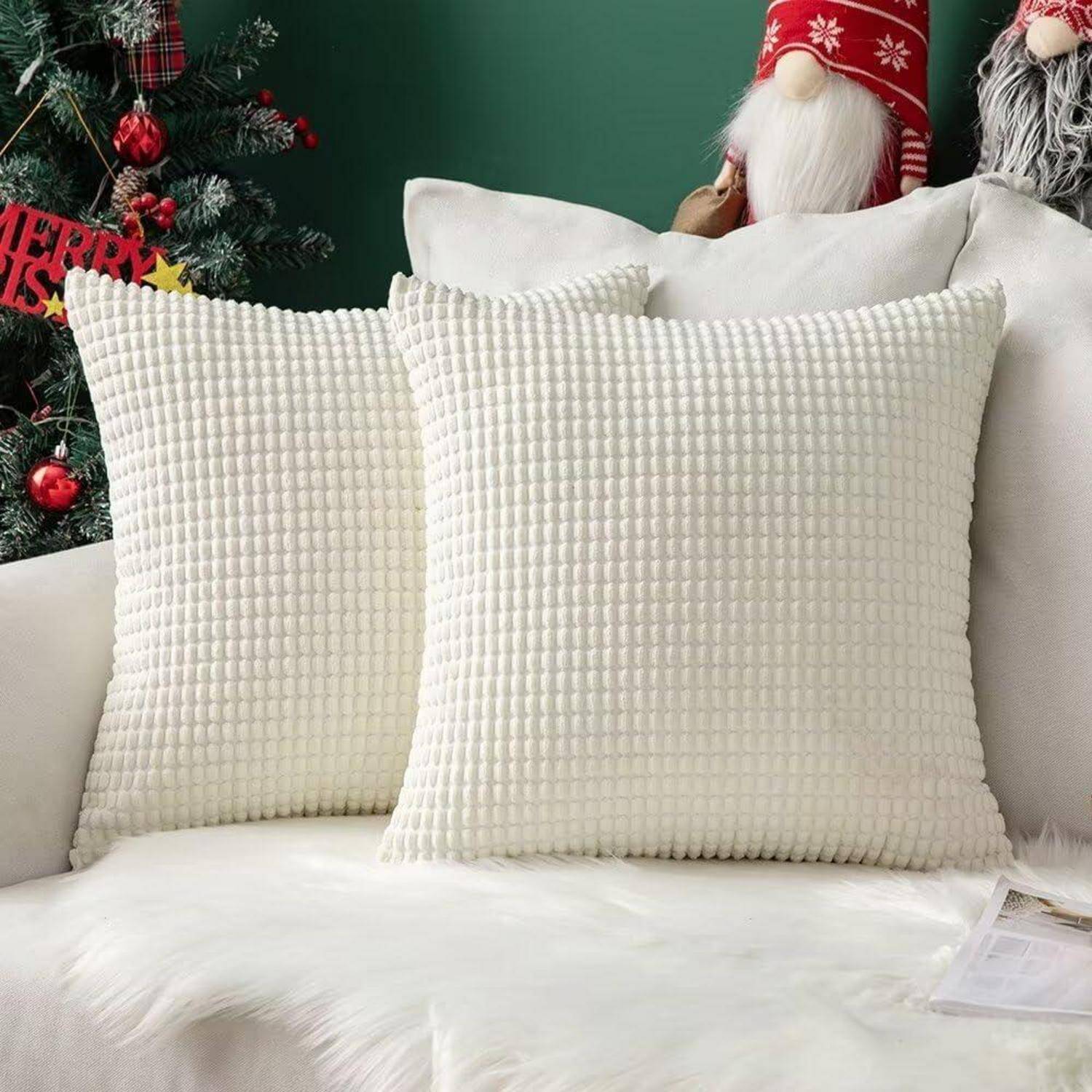 Zwei strukturierte weiße Kissen auf einem weißen Sofa, mit Weihnachtsdekoration und einer teilweise sichtbaren Weihnachtsmannfigur im Hintergrund.