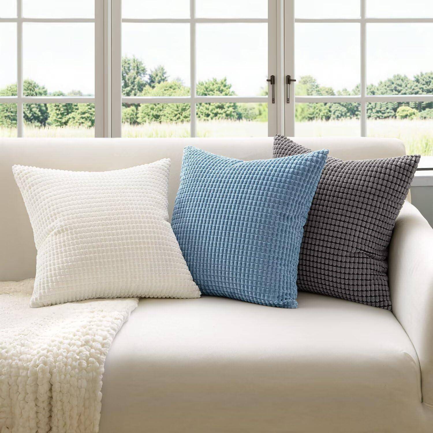 Ein weißes Sofa mit einer strukturierten Decke und drei Kissen: ein weißes, ein blaues und ein dunkelgraues. Große Fenster bieten Ausblick auf grüne Bäume.