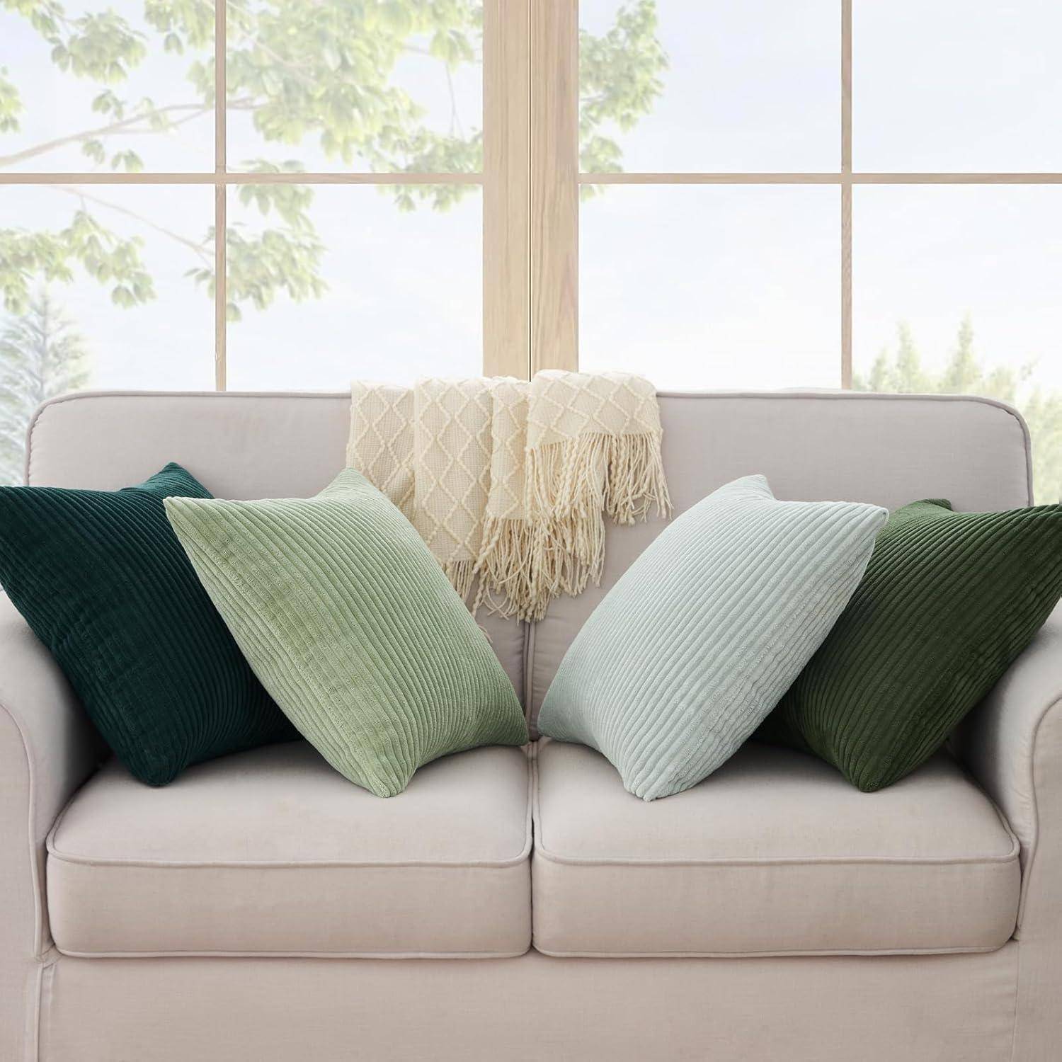 Ein beige Sofa mit vier gerippten Zierkissen in Grüntönen und einer cremefarbenen strukturierten Decke, platziert vor einem großen Fenster mit Blick auf Bäume draußen.