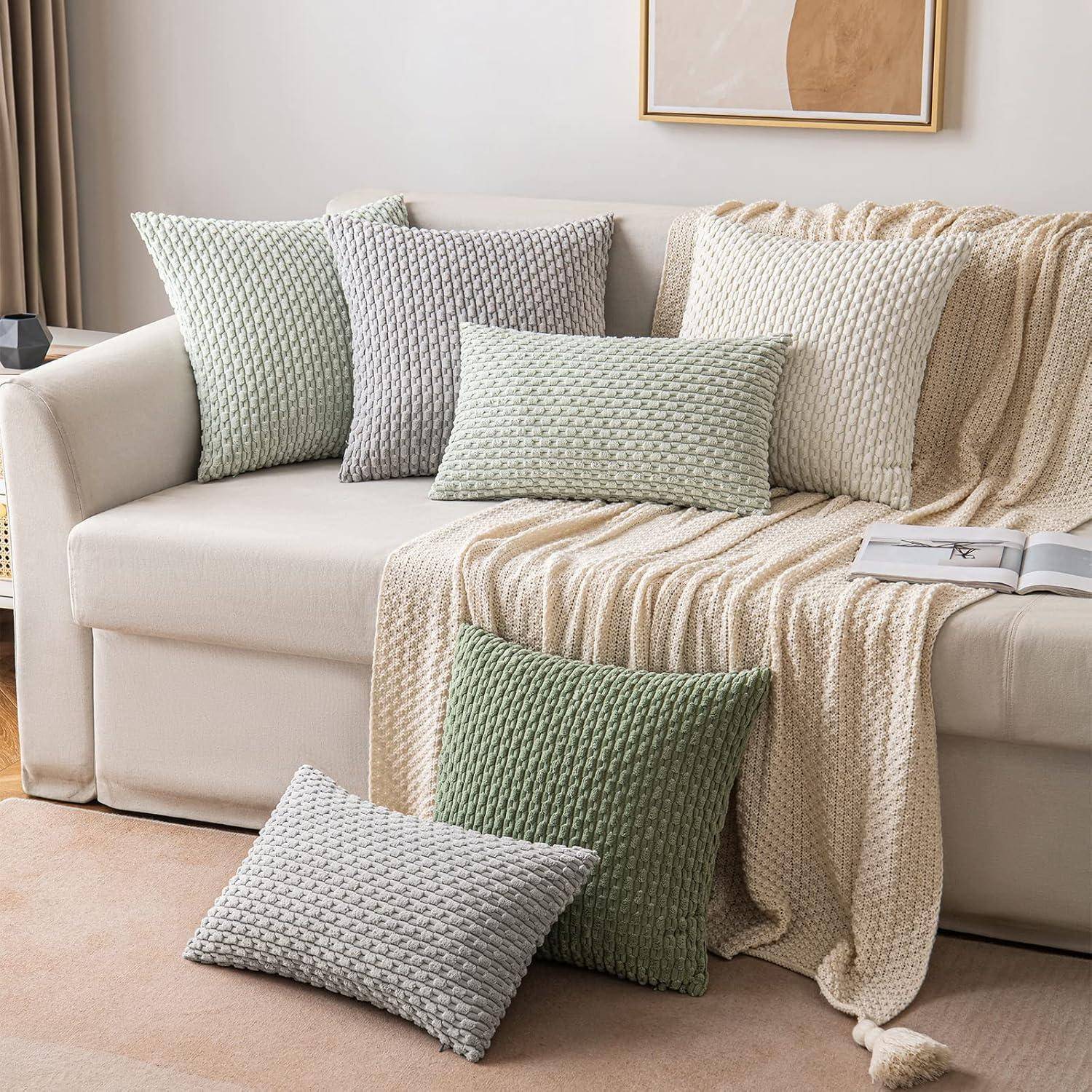 Ein weißes Sofa mit vier strukturierten Kissen in Grün- und Grautönen, eine beige Decke und ein gerahmtes abstraktes Kunstwerk an der Wand.