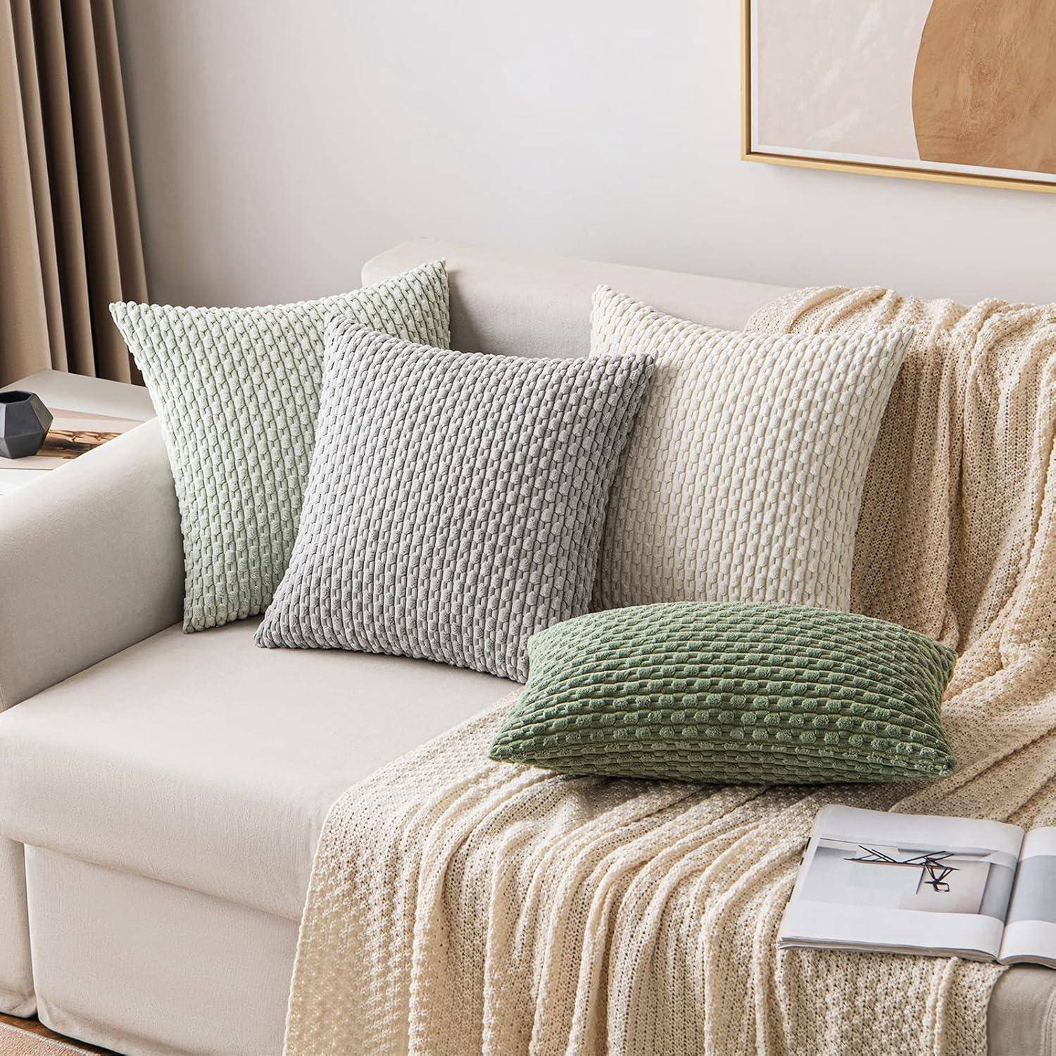 Sofa mit einer beigen Decke und drei strukturierten Kissen in Grau, Creme und Grün. Ein gerahmtes Kunstwerk ist teilweise an der Wand zu sehen.