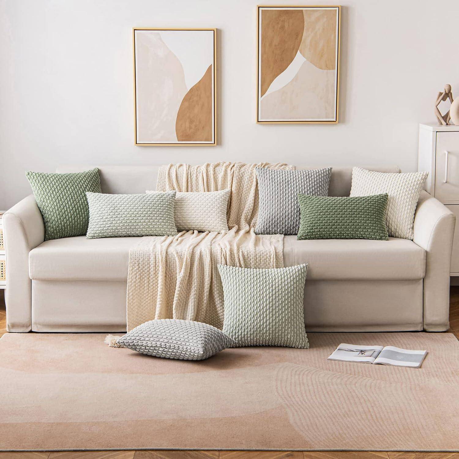 Ein beige Sofa mit pastellgrünen und cremefarbenen Zierkissen, einer passenden Decke und zwei abstrakten Wandkunstwerken. Ein Buch liegt aufgeschlagen auf einem Teppich.