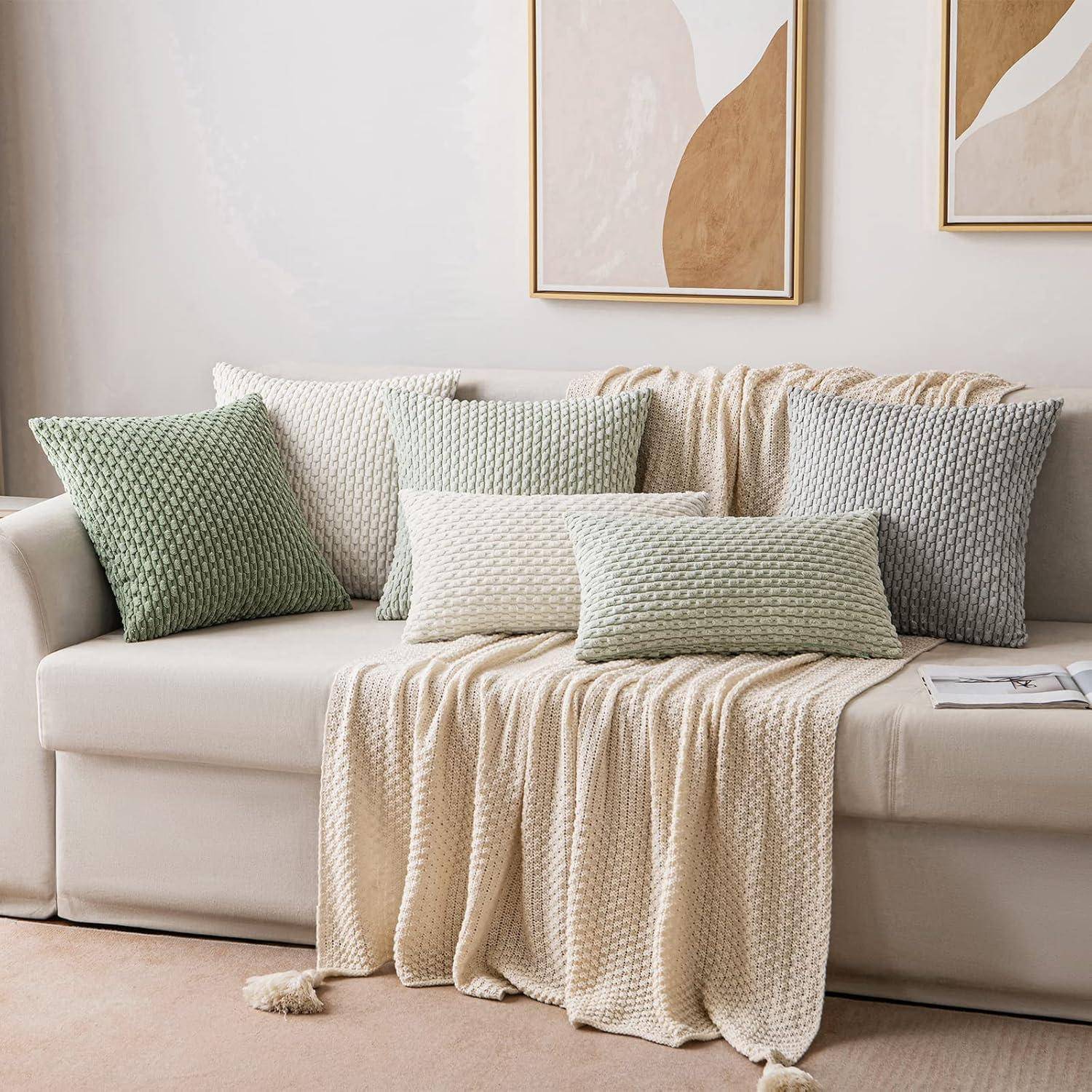 Ein beiger Sofa, geschmückt mit strukturierten Kissen in Grün-, Creme- und Weißtönen, überzogen mit einer passenden gemusterten Decke in einem minimalistischen Raum.