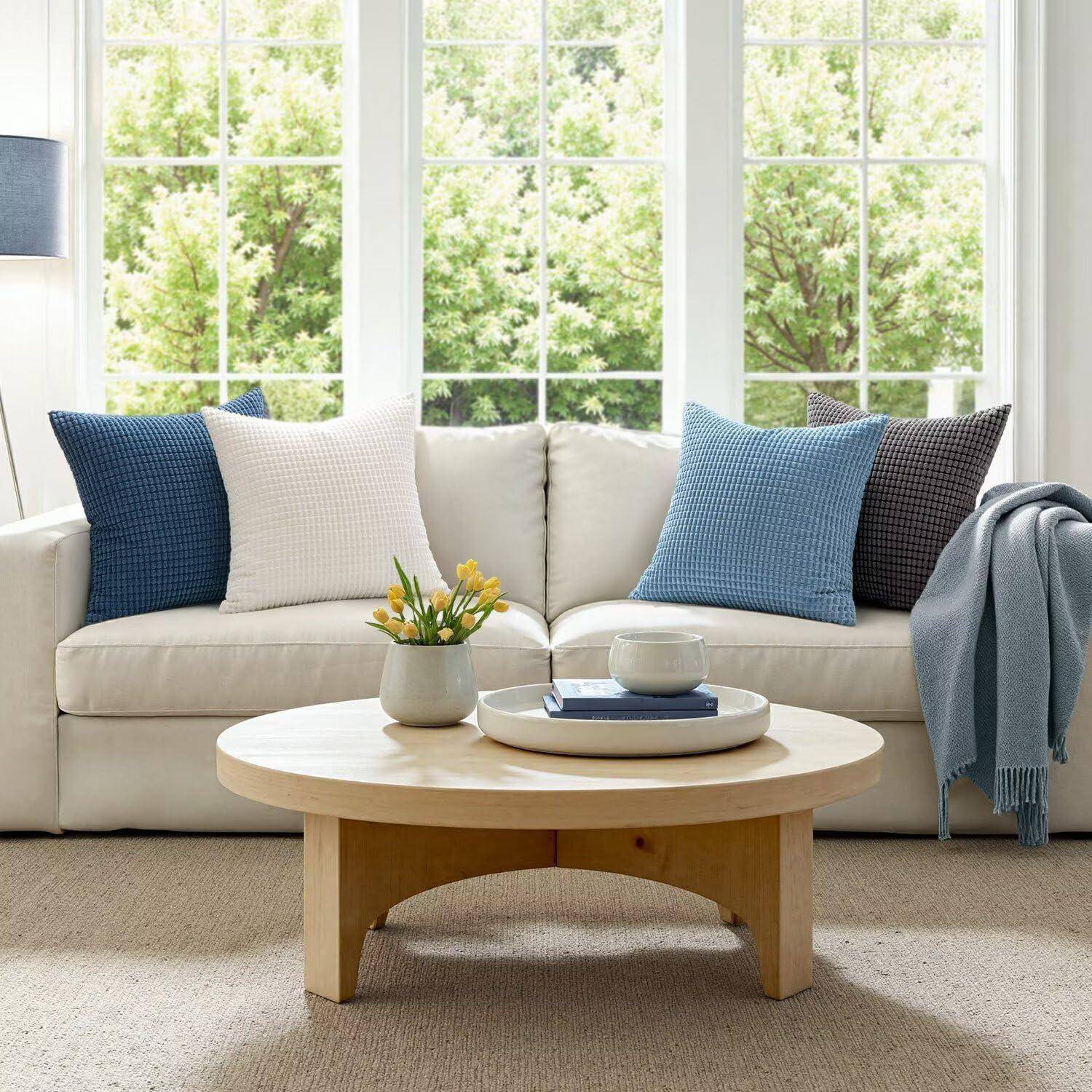 Gemütliches Wohnzimmer mit einem beigen Sofa, blauen und weißen Kissen, einem runden Holzcouchtisch und großen Fenstern, die Grünpflanzen im Außenbereich offenbaren.