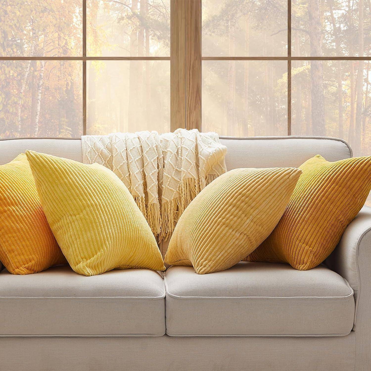 Ein gemütliches Wohnzimmer mit einem beigen Sofa, auf dem gelbe und orange Kissen liegen, eine gestrickte Decke darüber, und große Fenster, die einen sonnendurchfluteten Waldblick offenbaren.