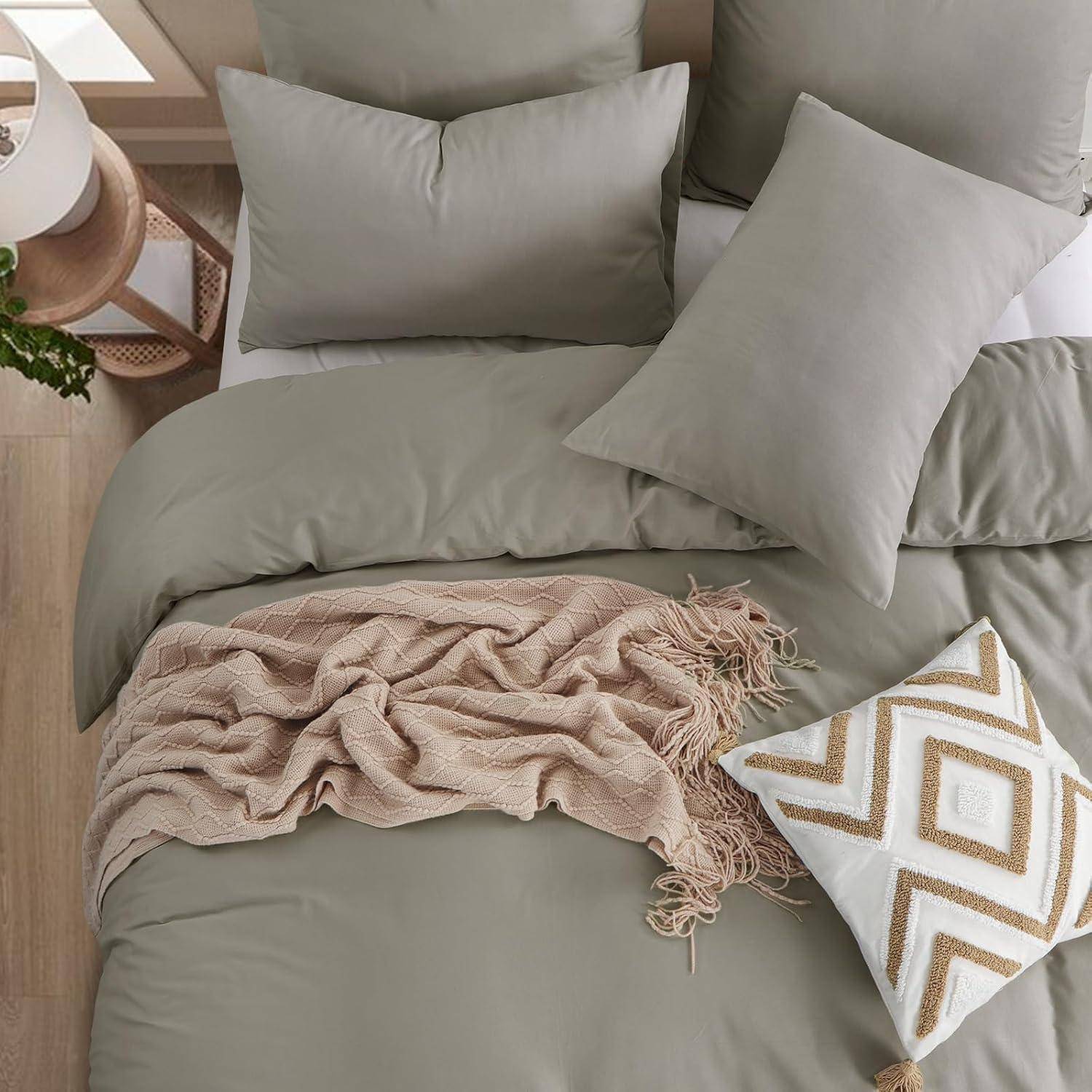 Ein ordentlich gemachtes Bett mit hellgrauen Laken, beigen und weißen Kissen und einer beigen Decke. Ein Holznachttisch trägt eine Topfpflanze.