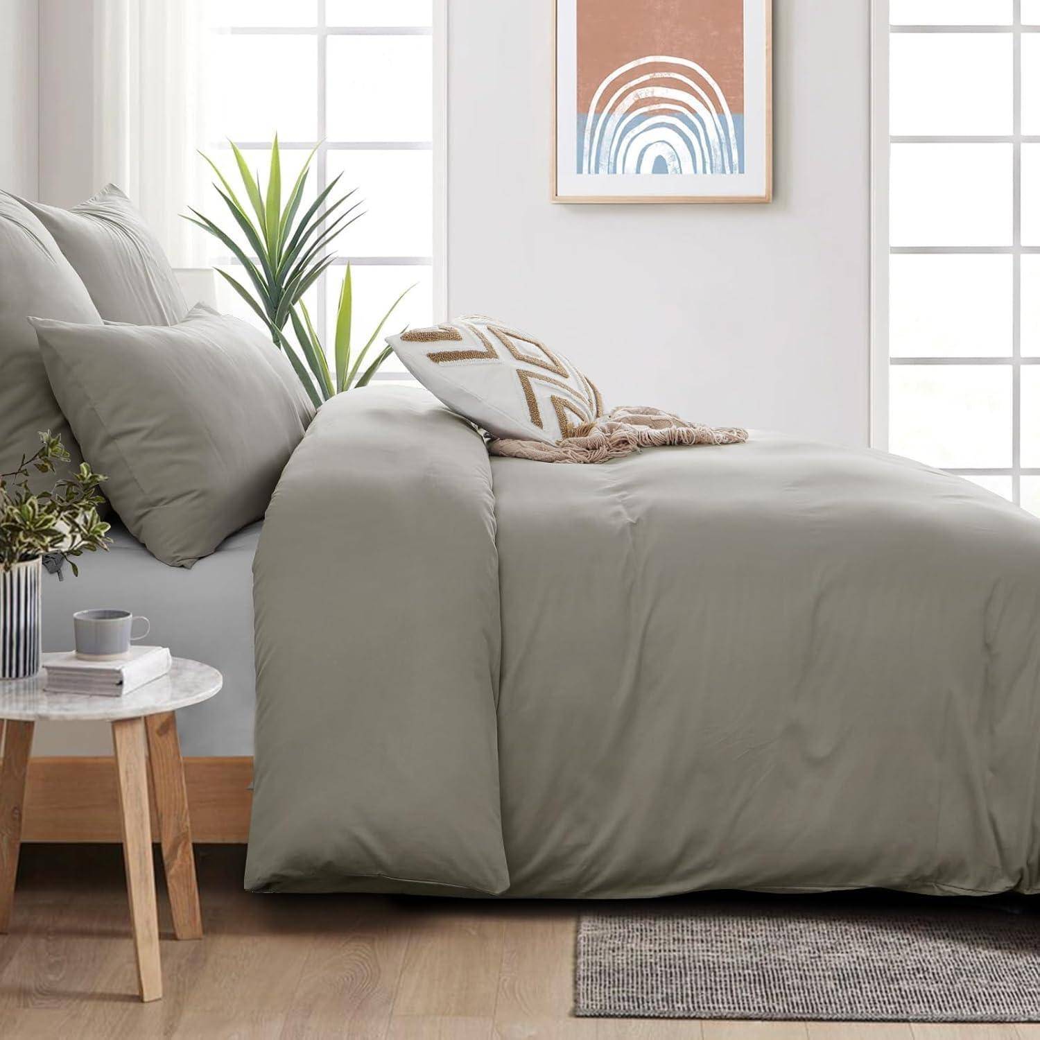 Ein ordentlich gemachtes Bett mit grauen Bettwäschestücken und gemusterten Kissen, neben einem kleinen Holz-Nachttisch mit einer Pflanze und einer Tasse, unter abstrakter Wandkunst.