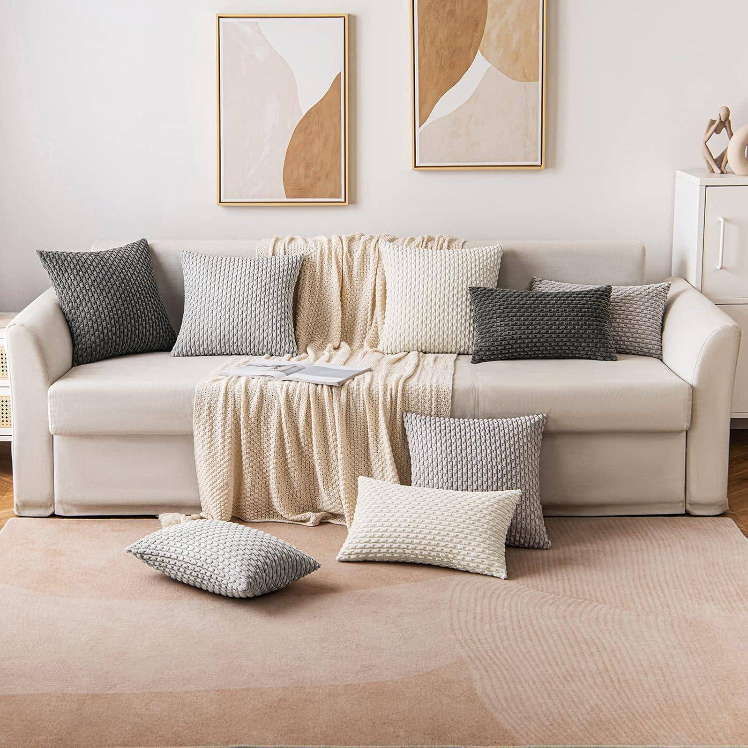 Ein moderner Wohnraum mit einem weißen Sofa, einer darüber gelegten Decke und sechs Kissen in Naturtönen mit Strukturmuster. Abstrakte Kunst an der Wand, die Eleganz verleiht.