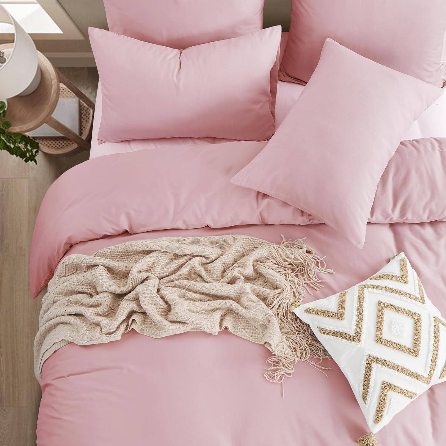 Ein ordentlich gemachtes Bett mit rosa Bettwäsche, rosa Kissen, einer beigen strukturierten Decke und einem weißen Zierkissen mit goldenen geometrischen Mustern.