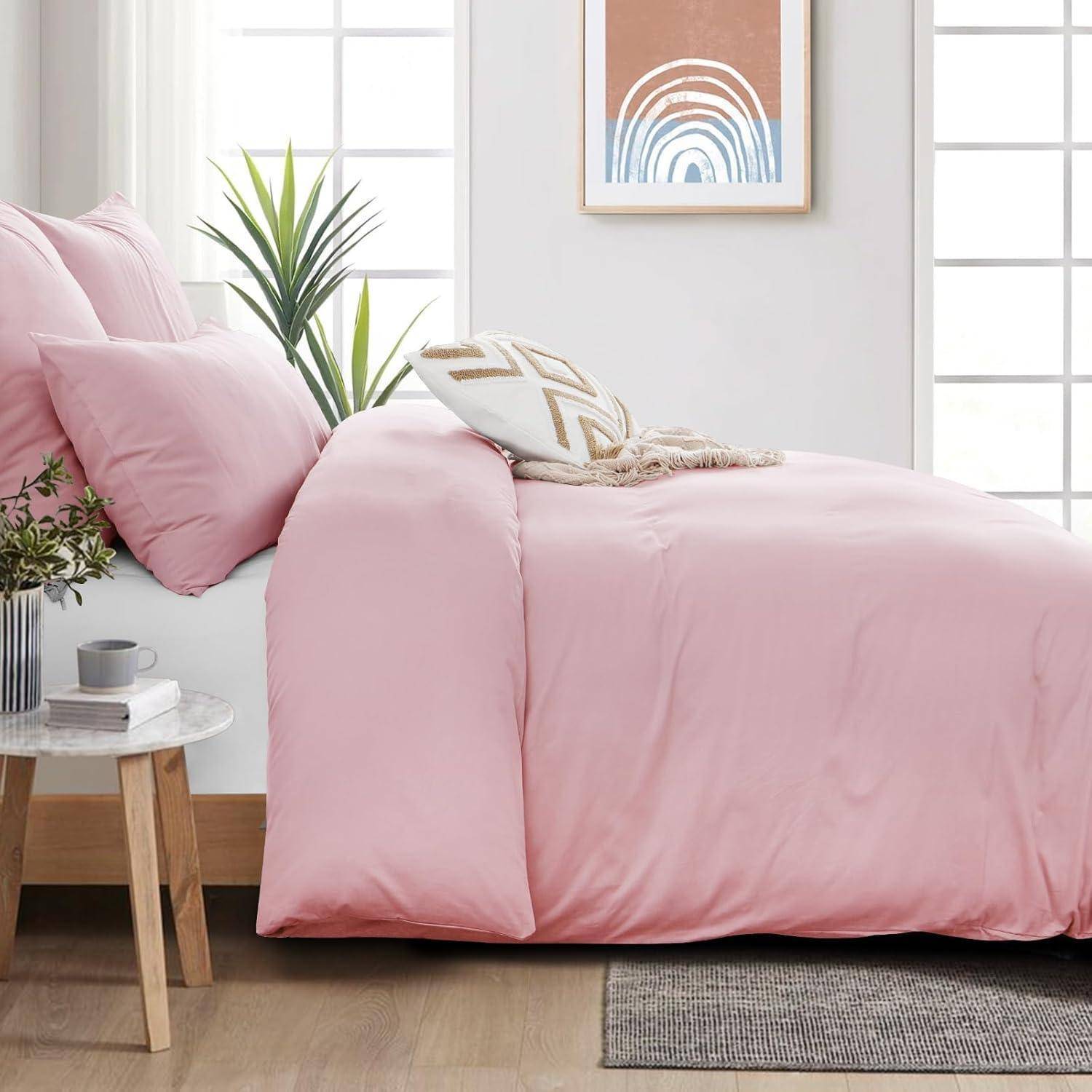 Ein Schlafzimmer mit weißen Wänden zeigt ein Bett mit rosa Bettwäsche und Kissen. Ein kleiner Nachttisch trägt eine Pflanze und eine Tasse. An der Wand hängt ein gerahmtes Kunstwerk.