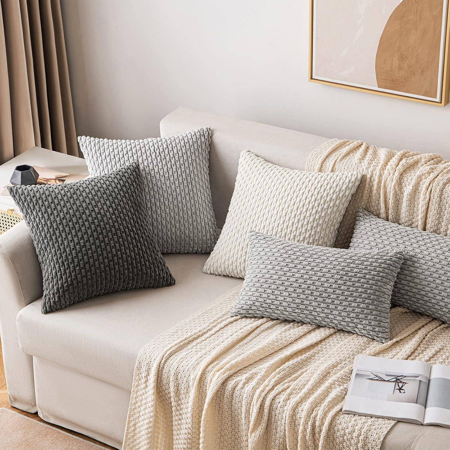 Eine gemütliche Wohnzimmerszene mit einem cremefarbenen Sofa, das mit strukturierten Kissen in Grau und Weiß verziert ist. Eine neutrale Decke liegt über der Armlehne des Sofas.