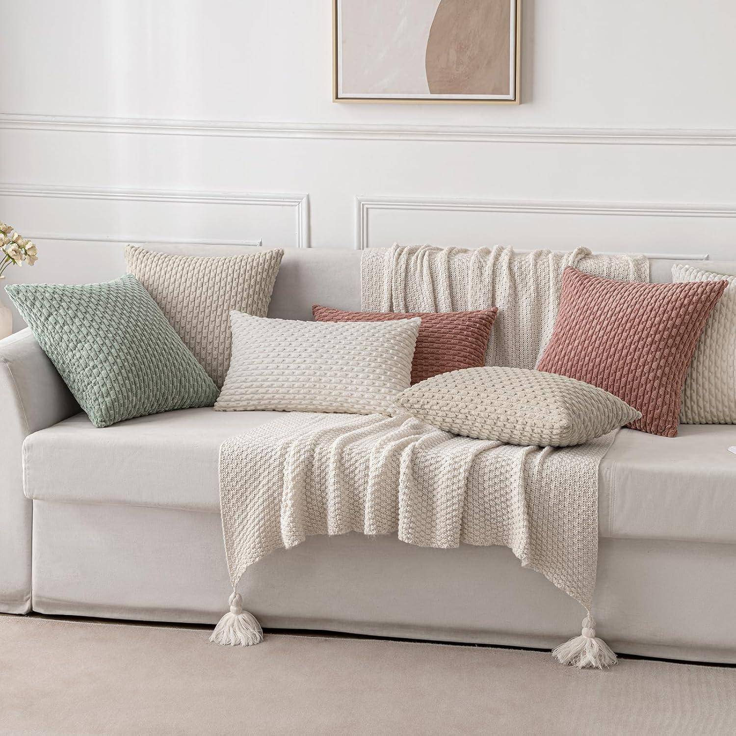 Ein weißes Sofa, geschmückt mit strukturierten Dekokissen in Pastellgrün, Beige und Terrakotta sowie einer passenden gestrickten Decke.