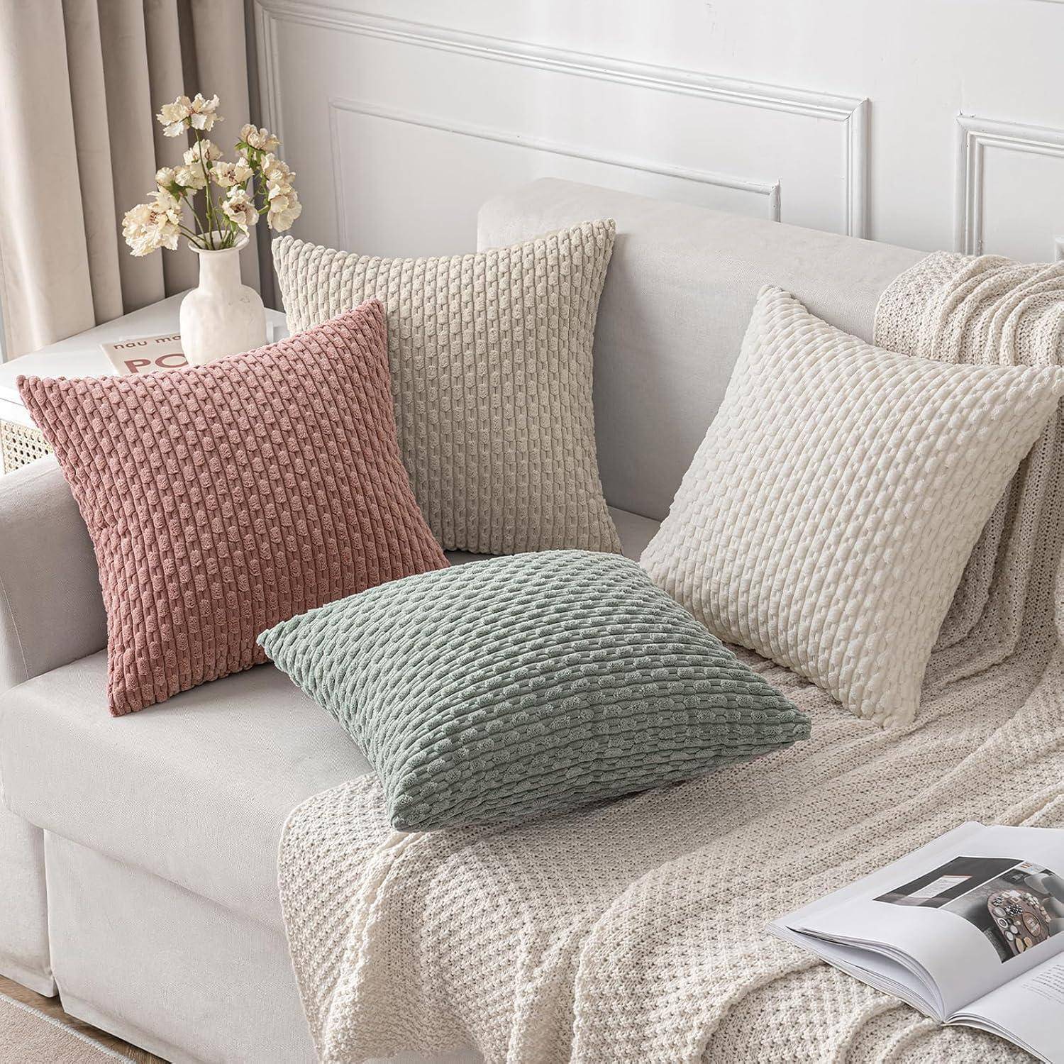 Eine gemütliche Wohnzimmerszene mit einem hellgefärbten Sofa, das mit strukturierten Kissen in Rosa, Creme und Grün geschmückt ist. Eine weiche Decke ist über die Armlehne gebreitet.