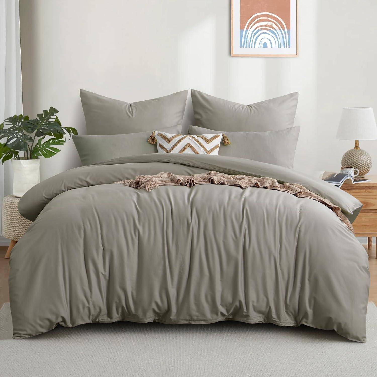 Ein ordentlich gemachtes Bett mit grauer Bettwäsche, passenden Kissen, einem gemusterten Zierkissen, flankiert von einer kleinen Lampe und einer Pflanze, die eine gemütliche Atmosphäre schafft.