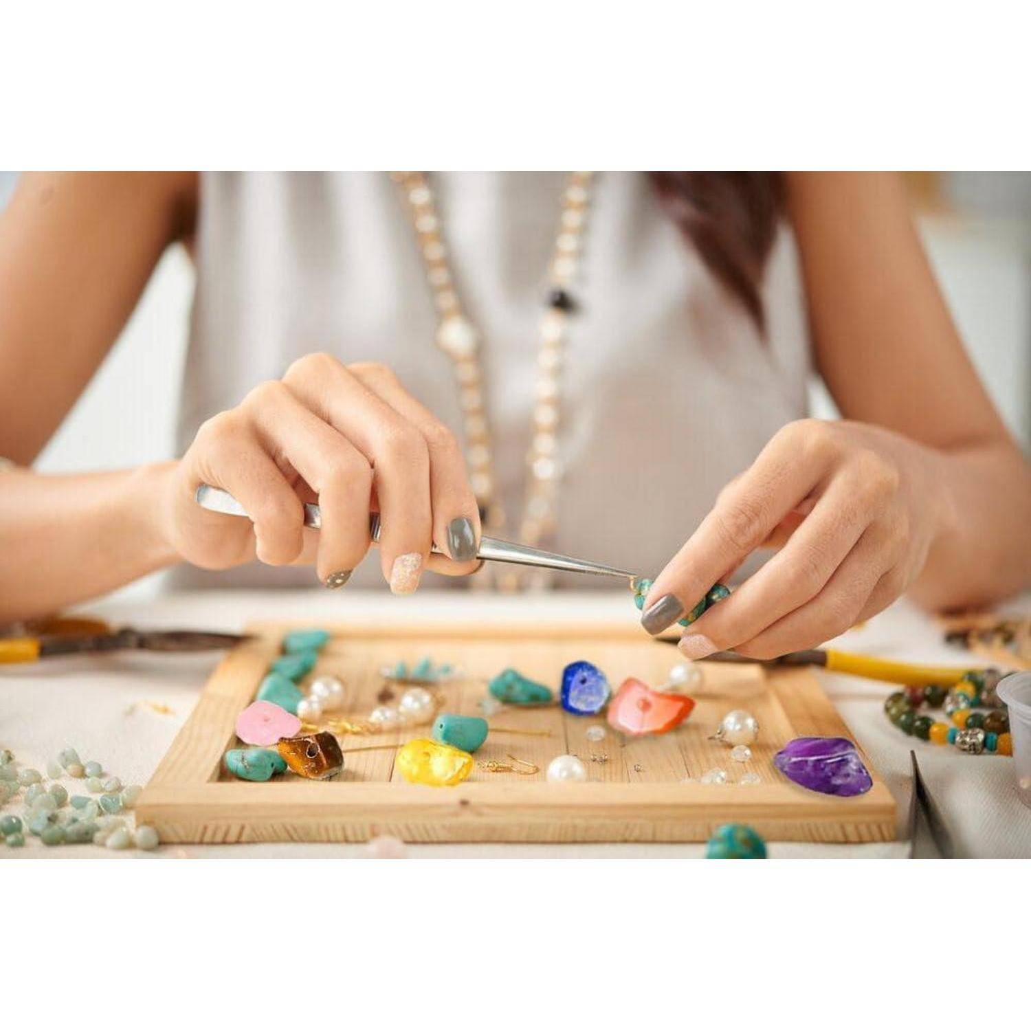 Eine Person stellt Schmuck her. Hände arrangieren bunte Perlen auf einem Holzbrett und verwenden dabei Werkzeuge. In der Nähe sind verstreute Perlen und Werkzeuge.