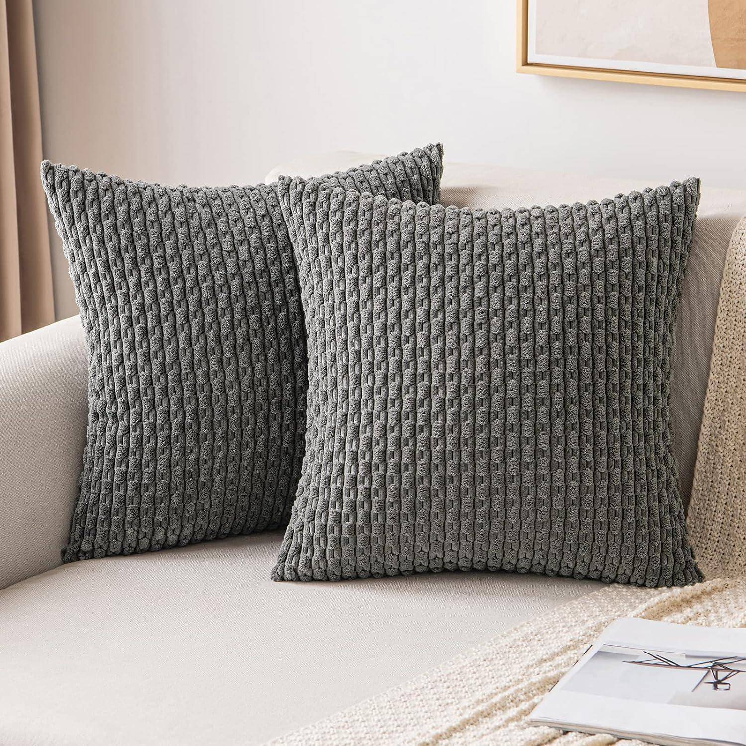 Zwei graue, strukturierte Kissen auf einem hellgefärbten Sofa mit einer gefransten Decke, die eine gemütliche und stilvolle Wohnzimmereinrichtung schaffen.