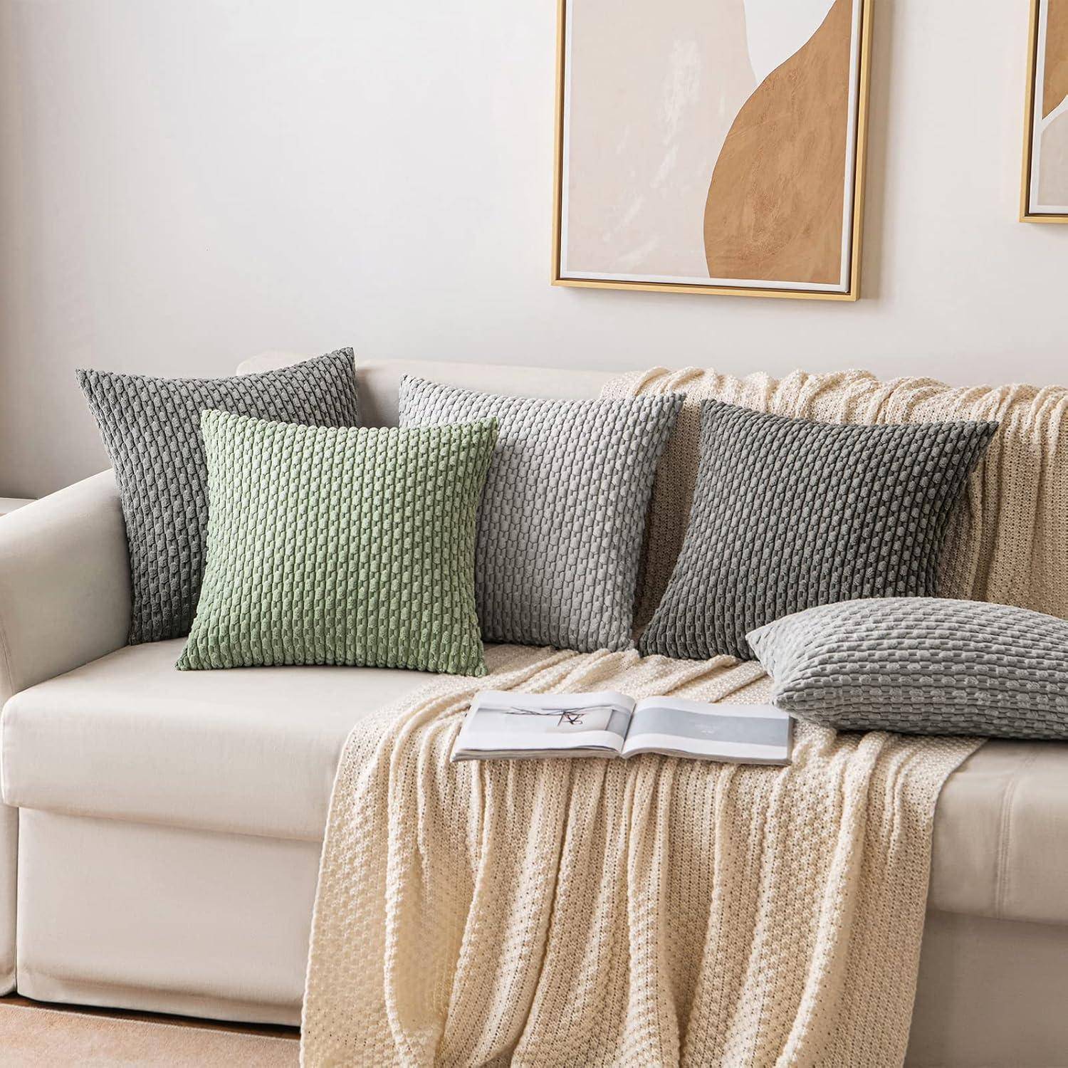 Ein gemütliches weißes Sofa mit strukturierten grauen und grünen Kissen, einer beigen gestrickten Decke und einem aufgeschlagenen Buch, vor einer abstrakten Wandkunst.
