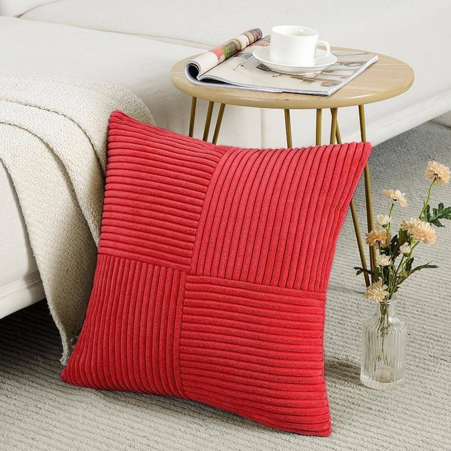 Ein rotes, quadratisches Kissen mit gestreifter Textur lehnt an einem Bett. Ein kleiner runder Tisch trägt eine Zeitschrift und eine weiße Tasse. In der Nähe befinden sich getrocknete Blumen in einer Vase.