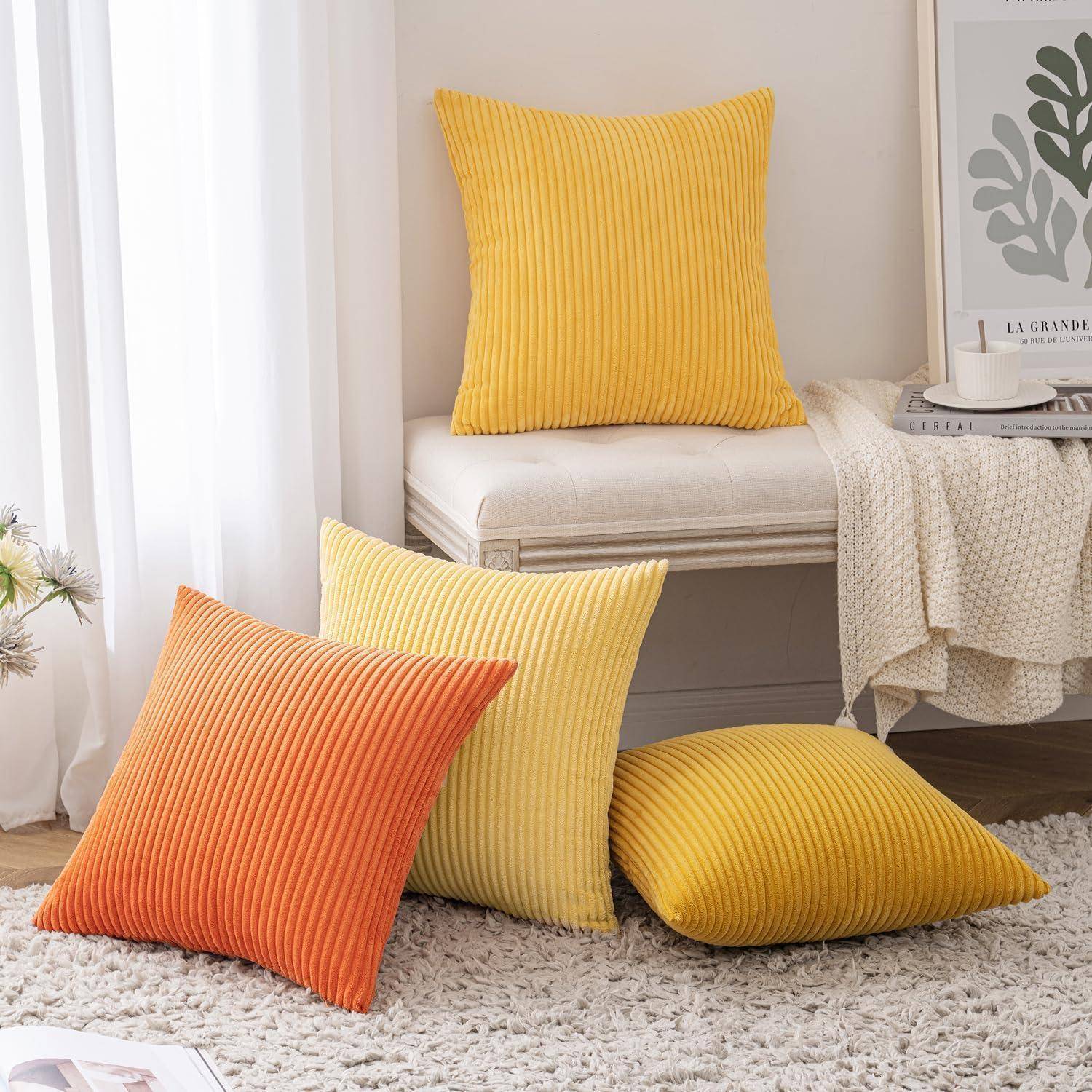 Drei gerippte Kissen in Orange und Gelb sind auf einem weißen Sofa und Boden angeordnet. Eine gefaltete Decke und eine Pflanze befinden sich im Hintergrund.